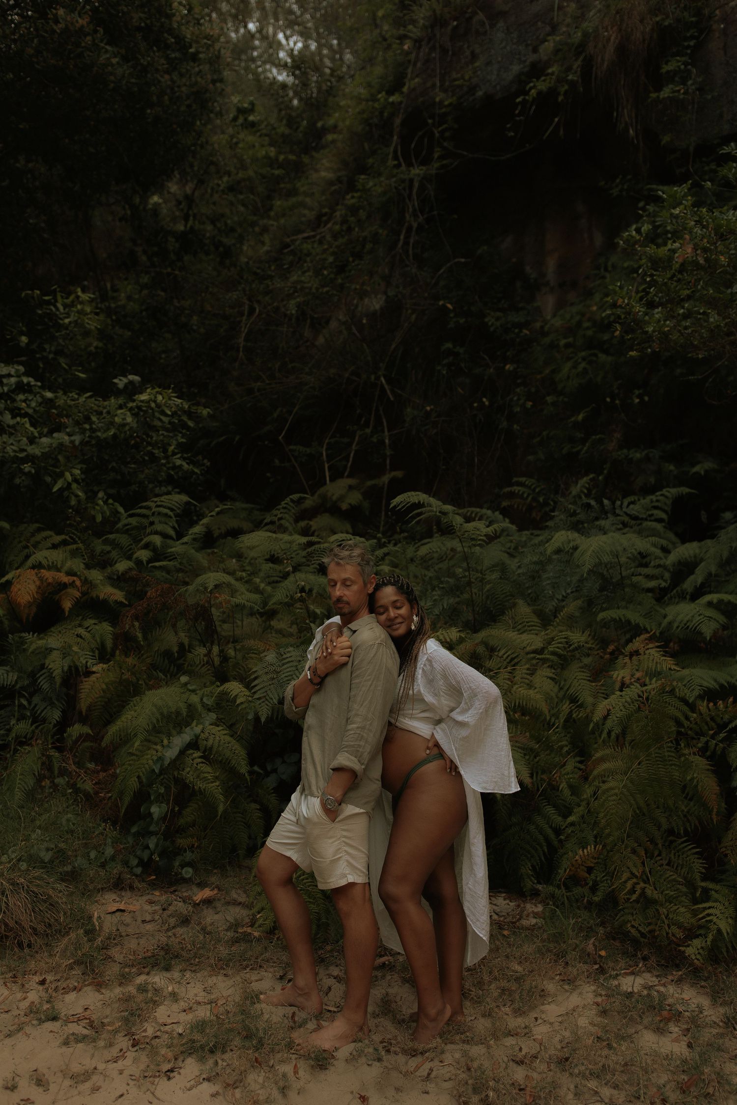 Two figures embrace on a rocky outcrop surrounded by dense ferns and forest vegetation in moody lighting.