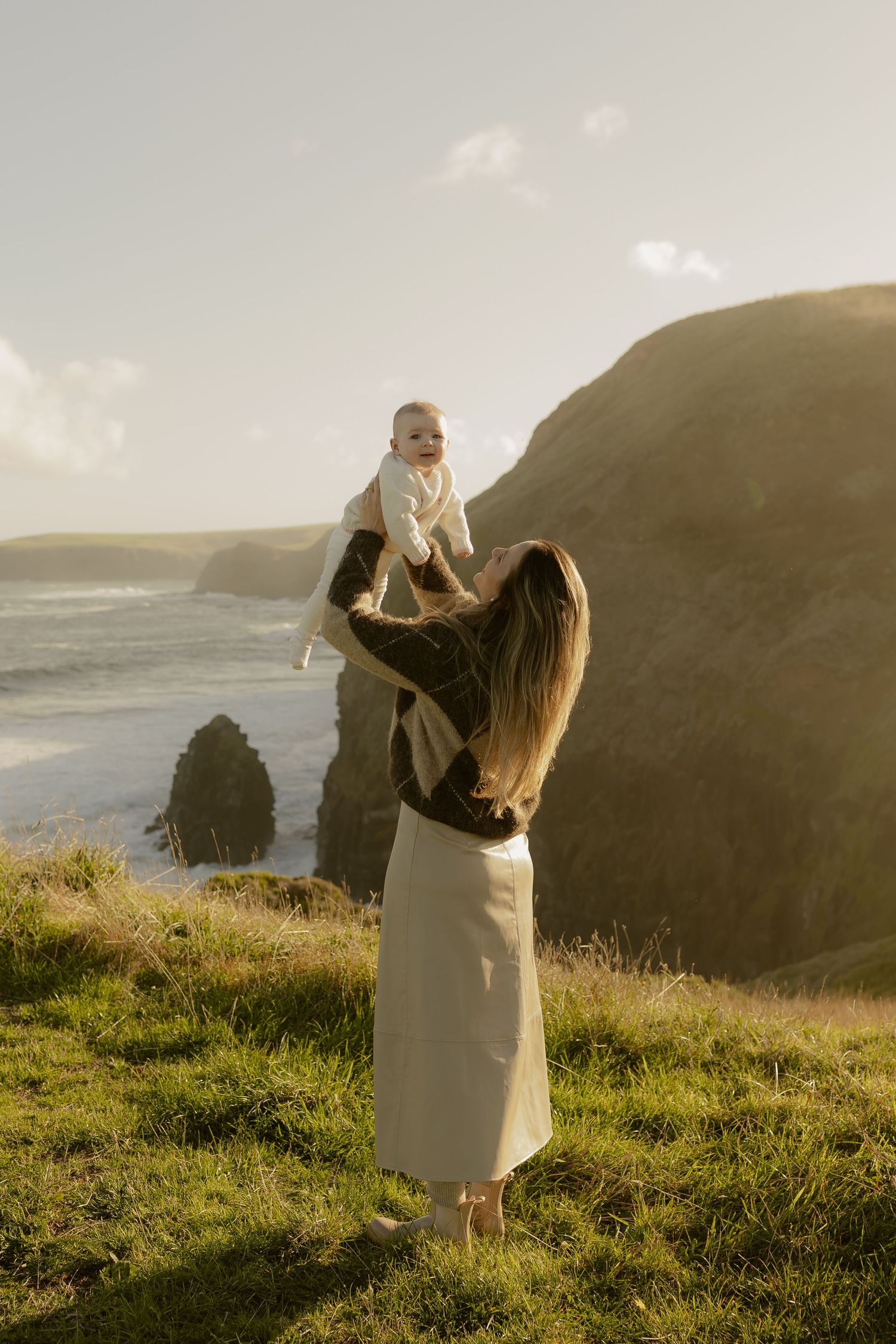Mother lifting baby above coastal cliffs at golden hour.
