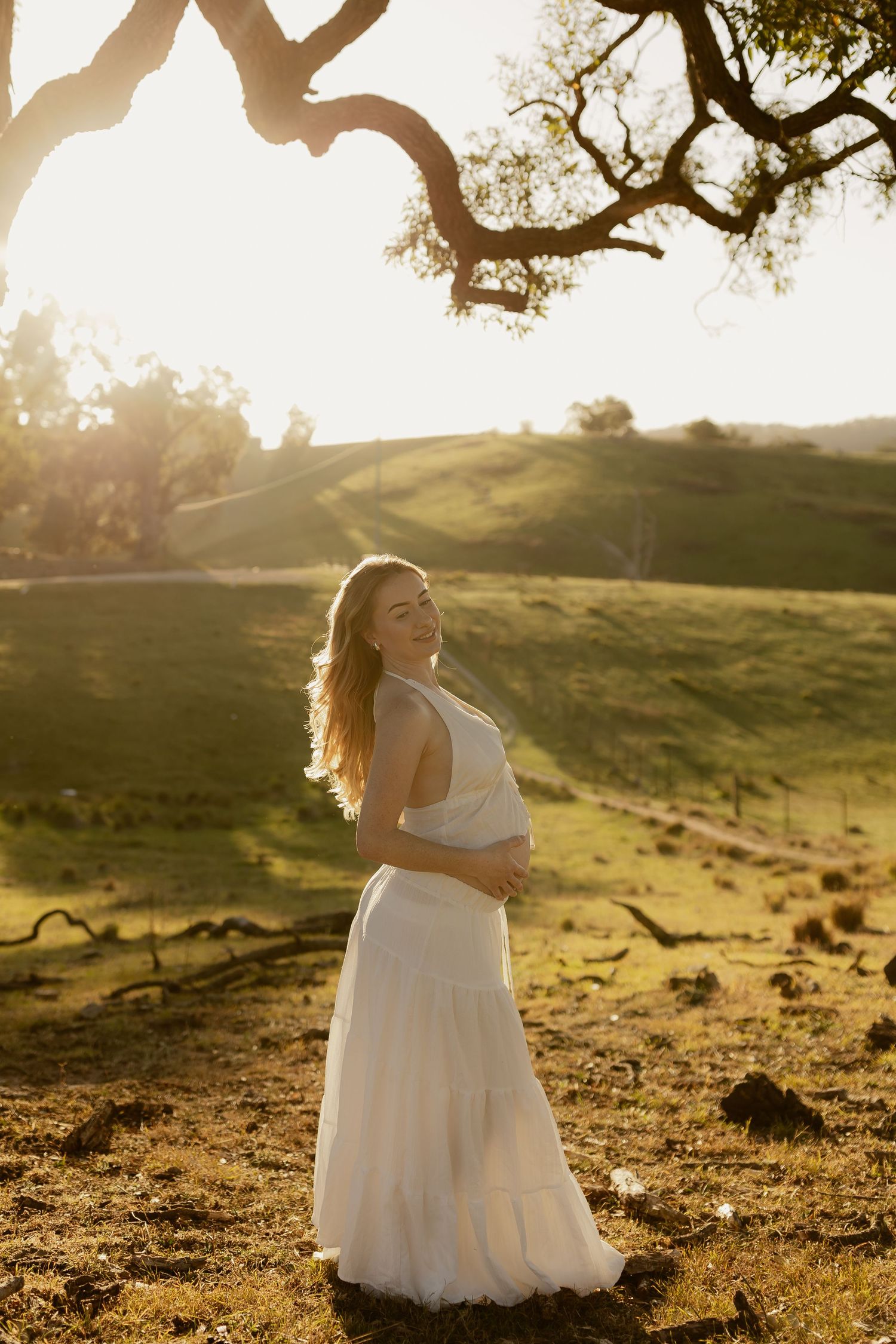 Pregnant woman in white dress holding baby bump, standing in sunlit green field under oak tree.