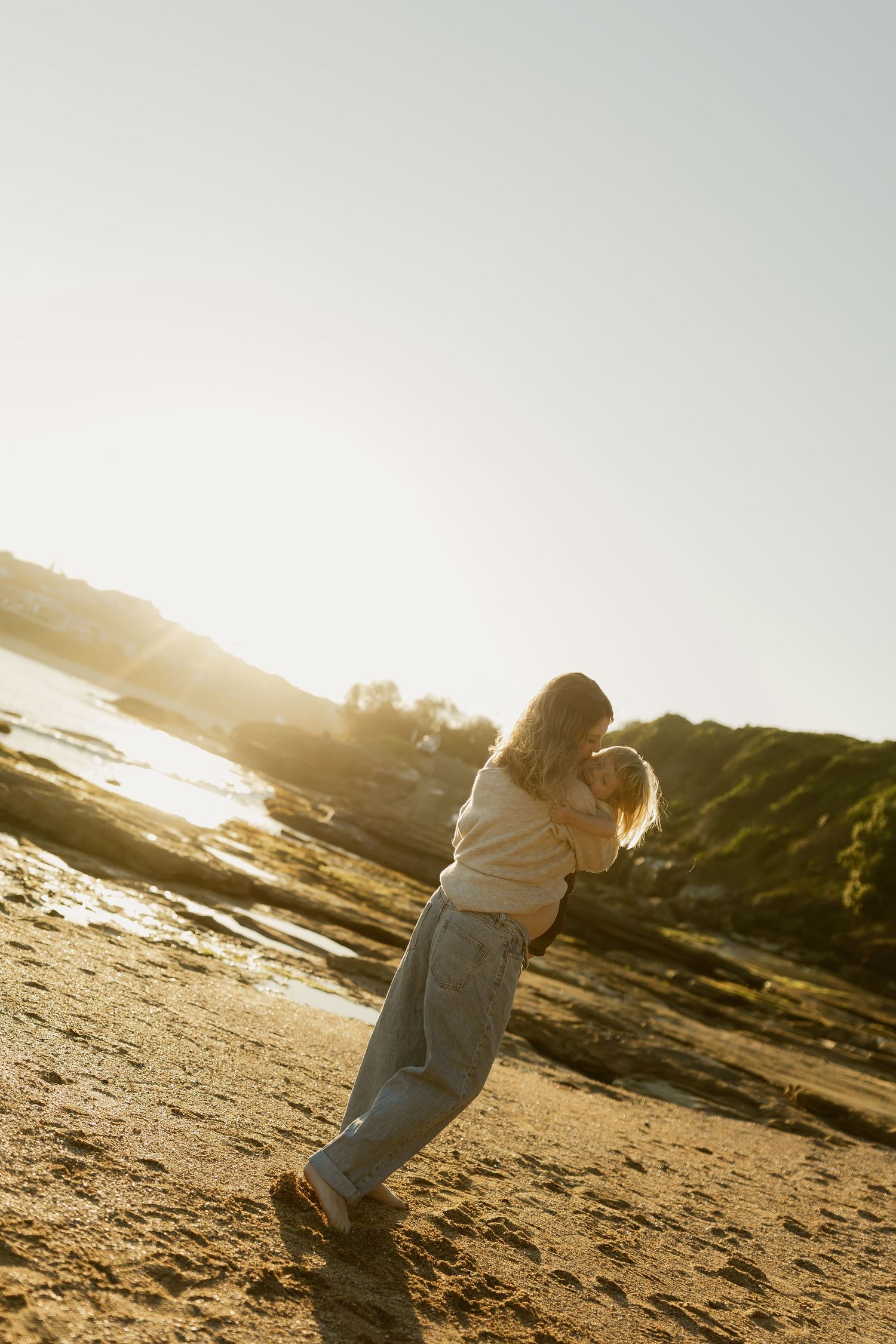 Woman carrying a child on a sandy beach at golden hour sunset with rocky coastline in background.