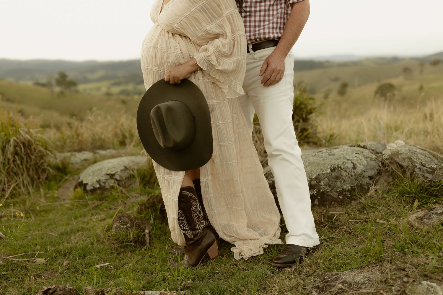 Couple embracing outdoors on grassy hill, woman in cream dress holding cowboy hat, man in plaid shirt and white jeans.