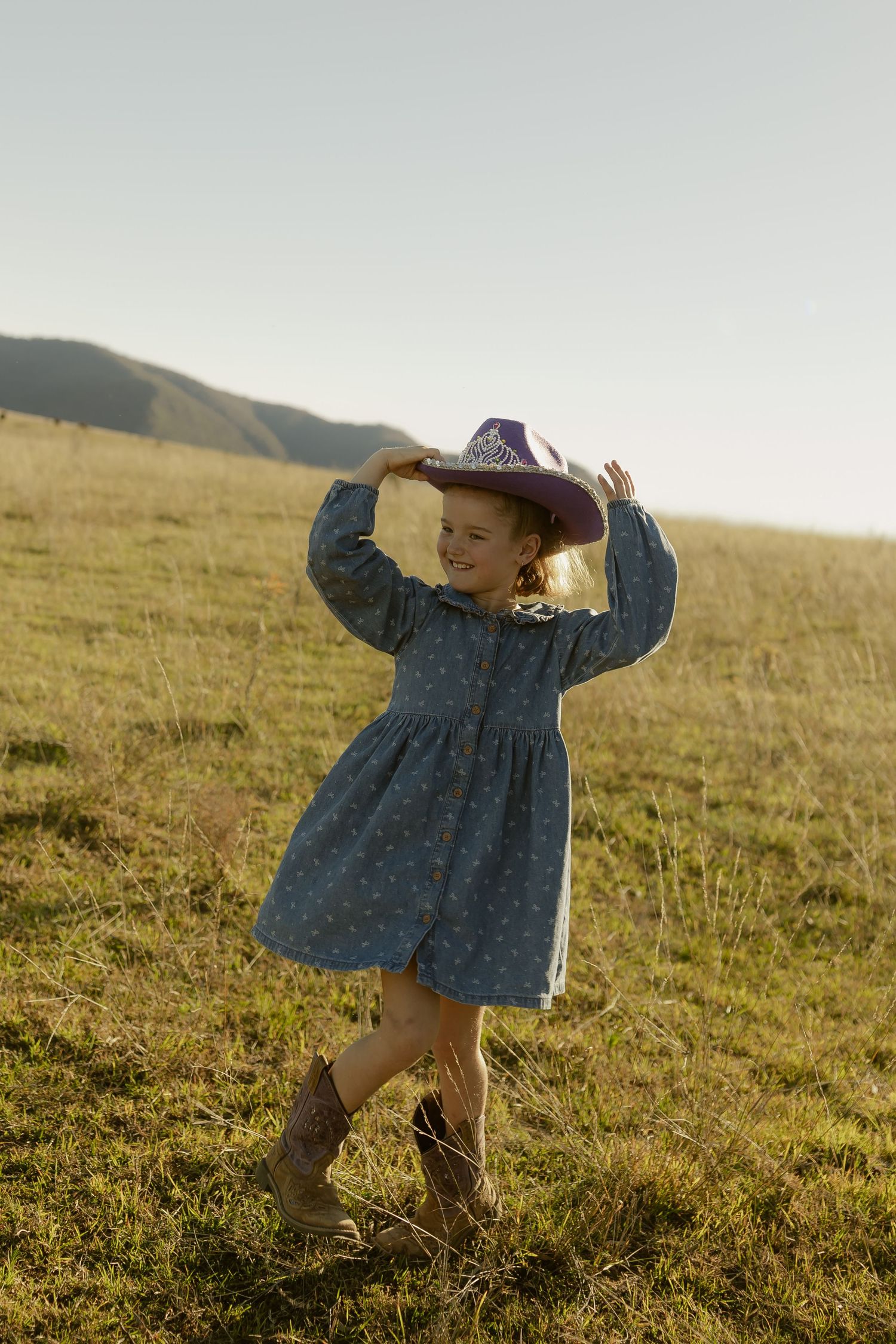 A young girl in a blue polka dot dress and cowboy boots holds a purple cowboy hat on her head in a sunny field.