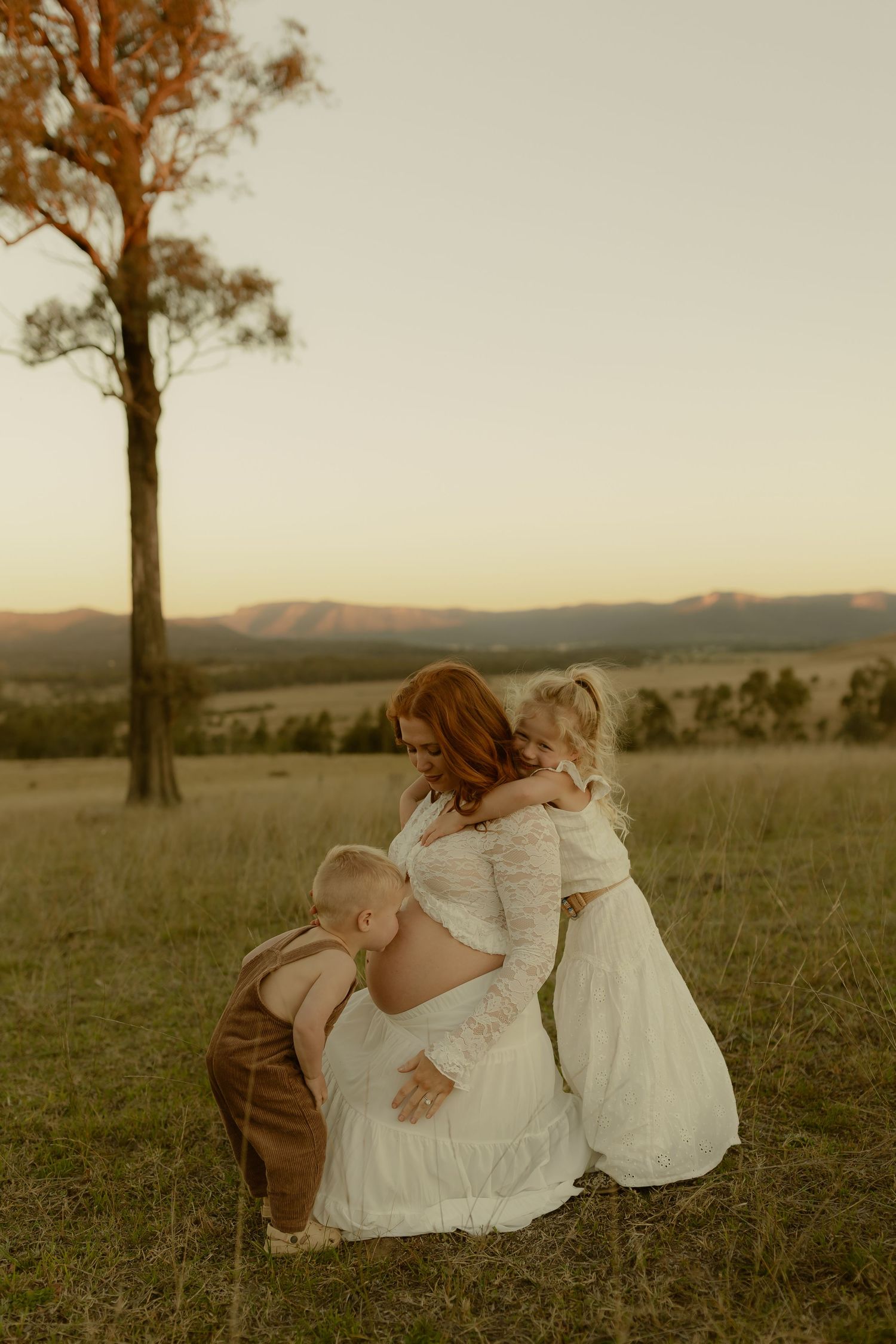 Pregnant mother crouching to embrace two young children in sun-drenched field at dusk.