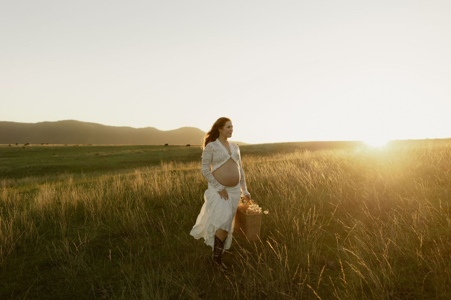 Pregnant woman in white dress holding basket in golden sunset field with mountain backdrop.