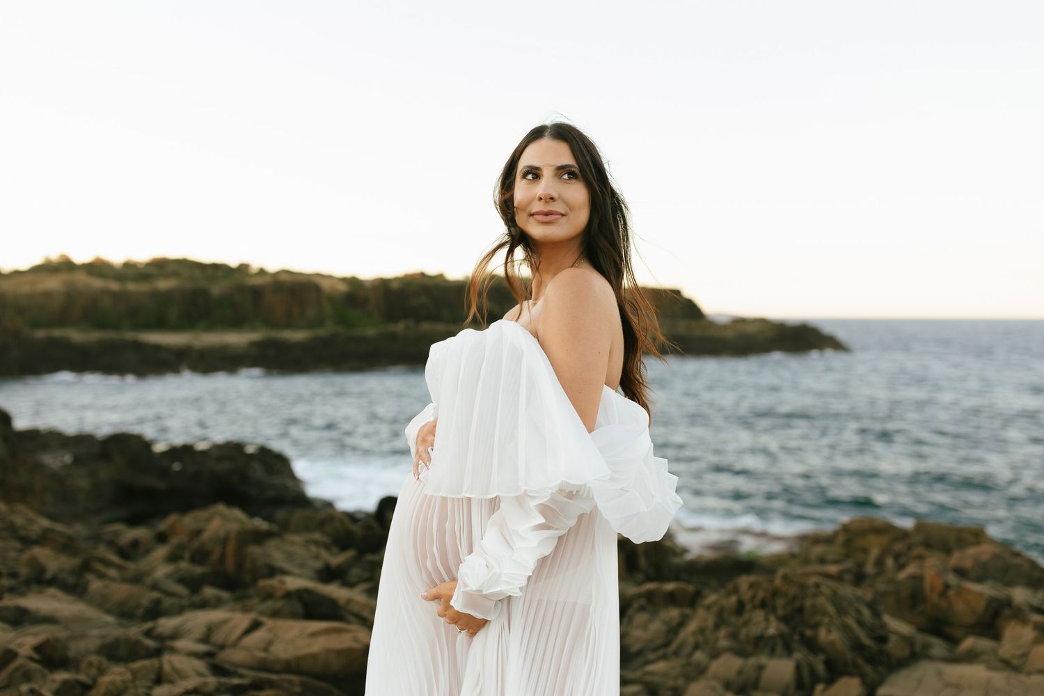 Elegant maternity portrait of woman in white dress against rocky coastal landscape at sunset.