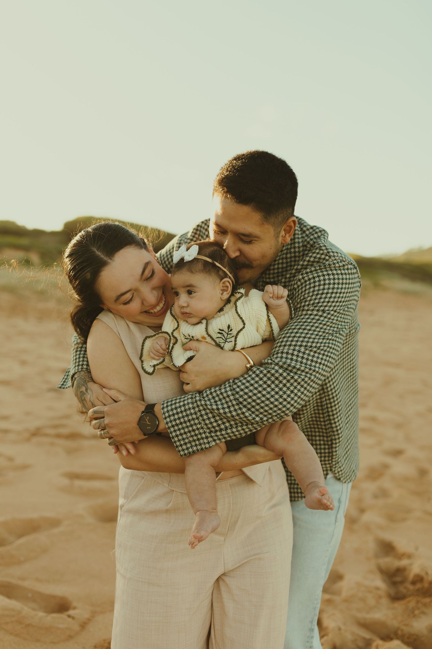 A family shares tender embraces while spending time together on sandy dunes at sunset in matching neutral tones.