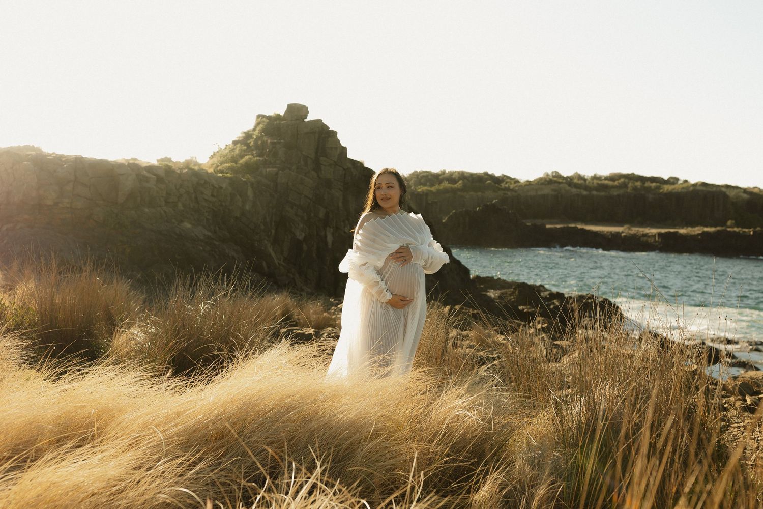 Ethereal maternity photograph capturing a glowing moment at a coastal cliff during sunset.