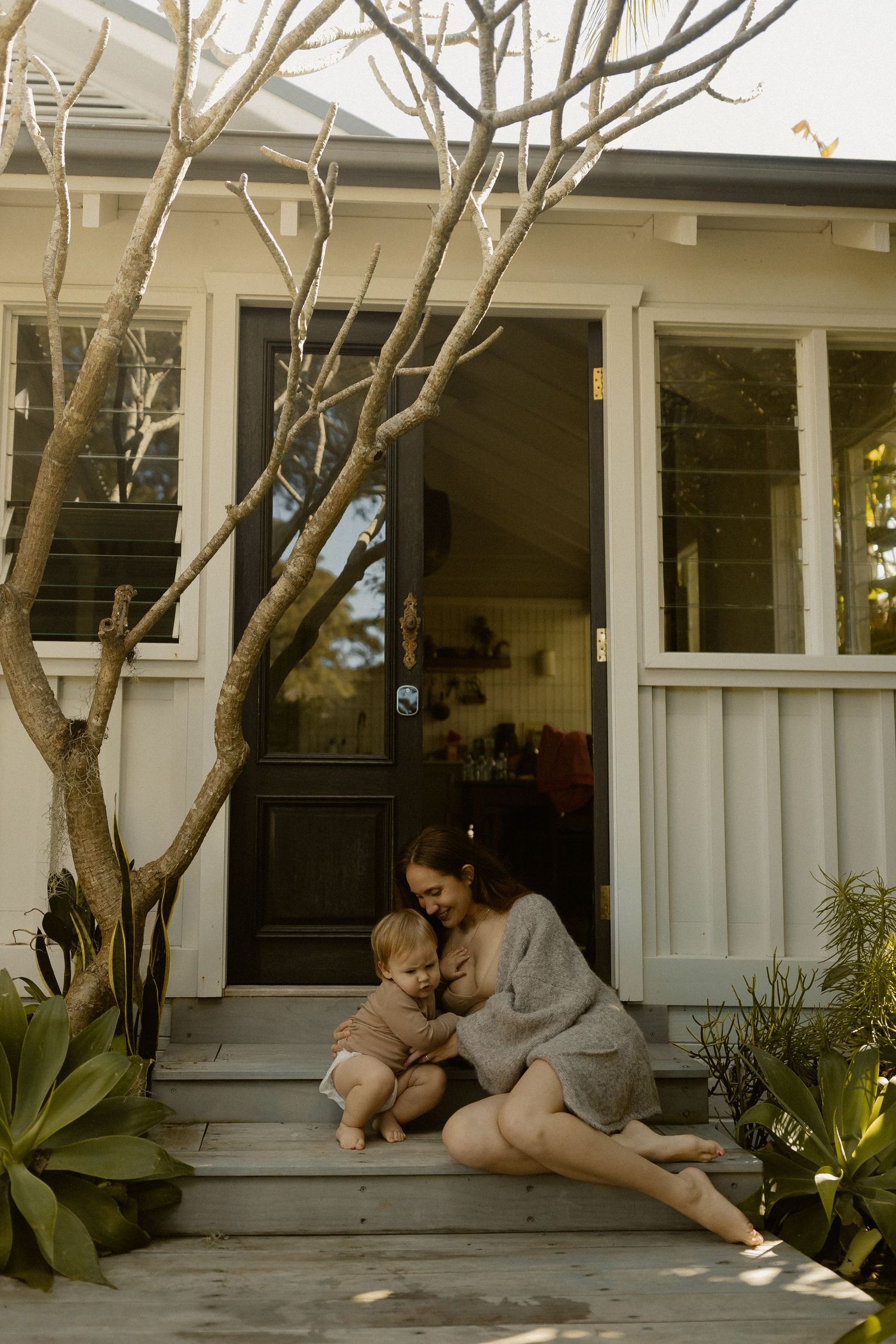 Mother and baby sharing tender moment on front porch of craftsman style home at sunset.