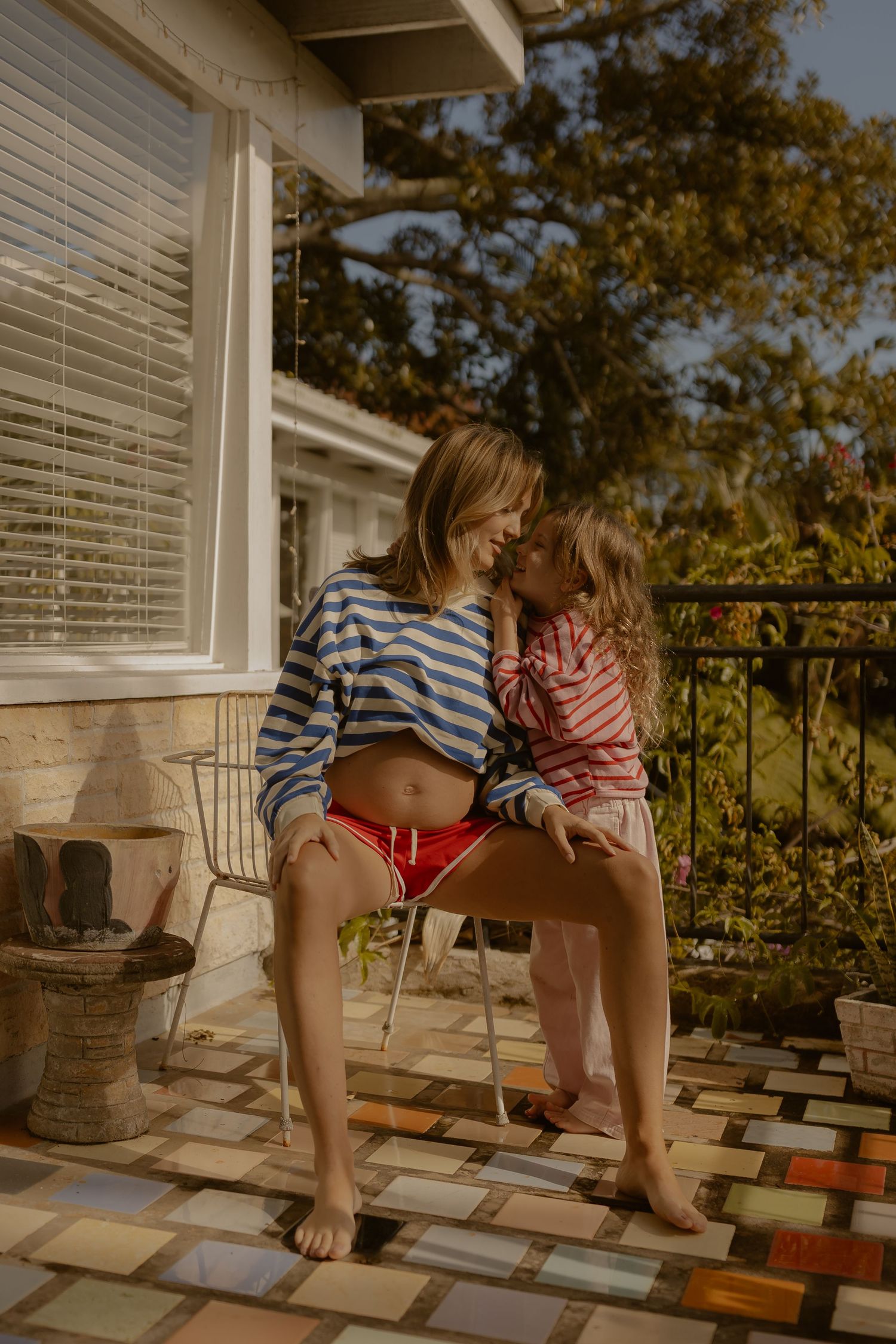 Pregnant mom sitting on porch chair holding toddler daughter on sunny day.