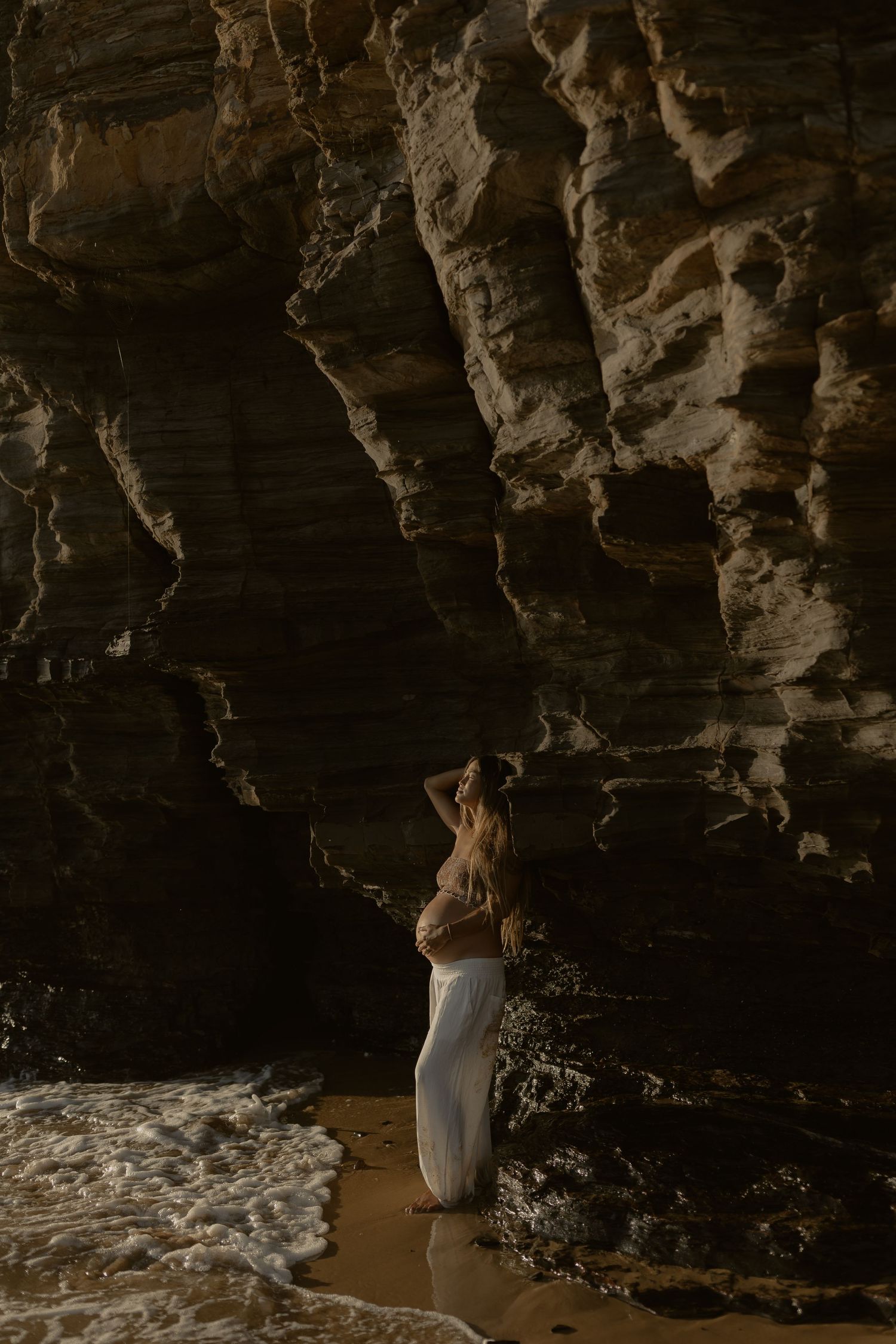 Pregnant woman in white skirt stands against dramatic rocky cave wall on sandy beach with ocean waves.