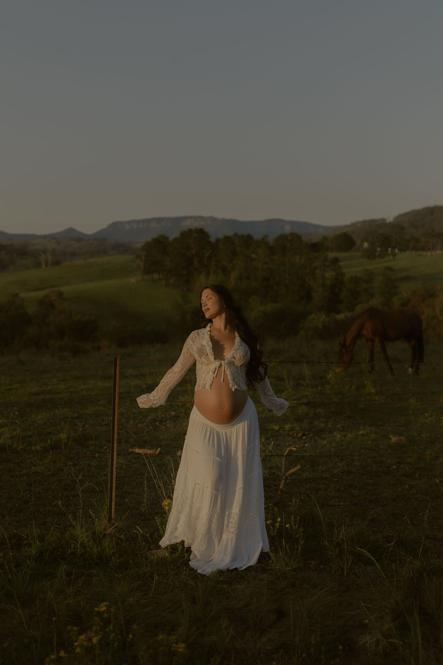 Pregnant woman in white lace dress standing in green field at golden hour with horse grazing and rolling hills behind her.