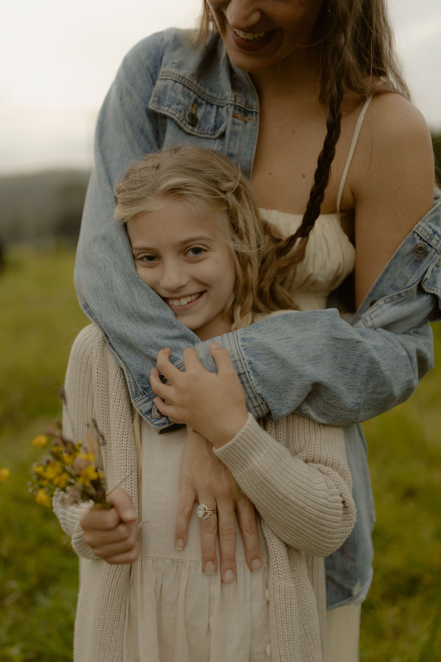 A smiling girl holding wildflowers is embraced by a woman in a denim jacket in a green field.