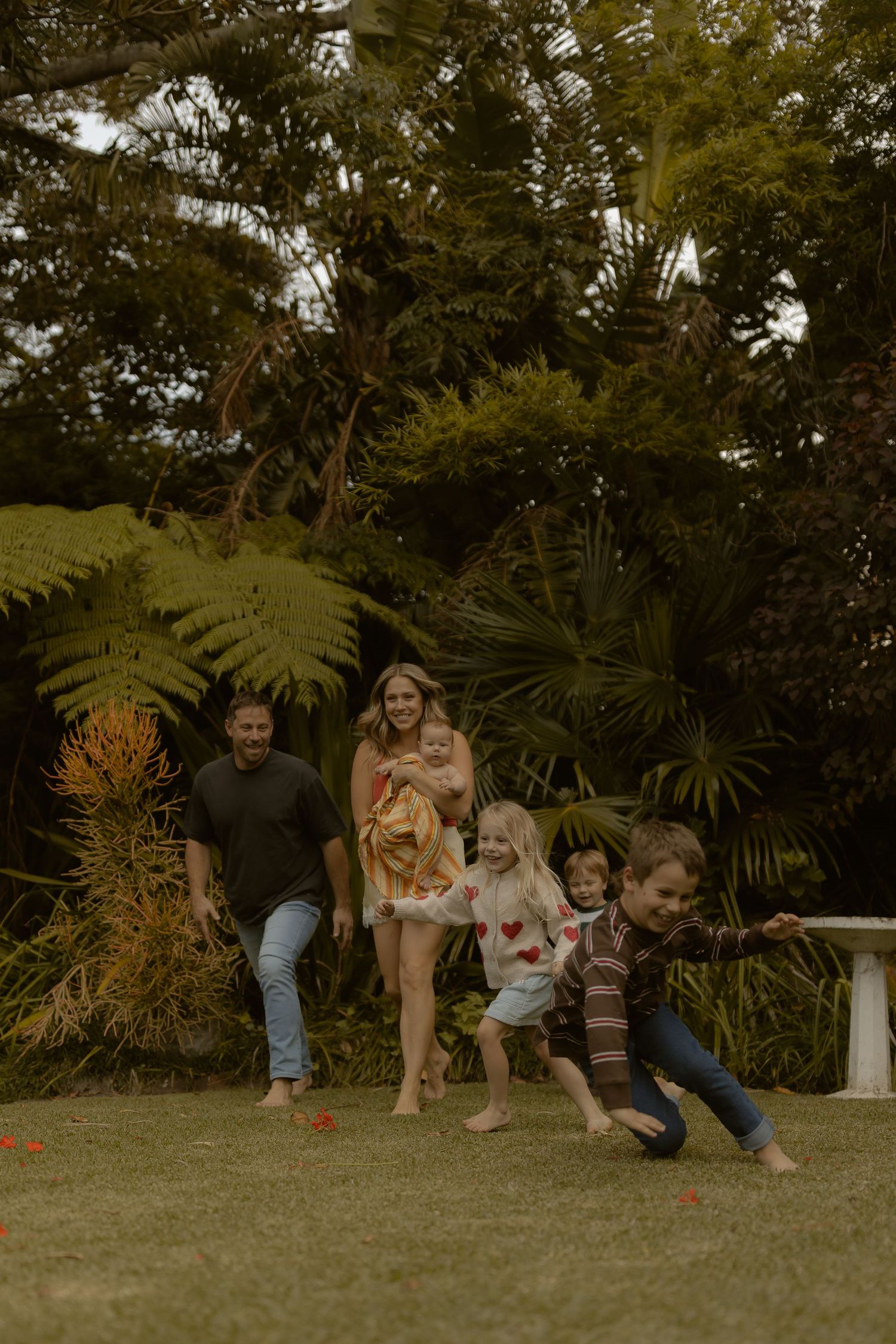 Family playing together in a lush green garden, parents and children laughing outdoors on grass surrounded by tropical ferns.