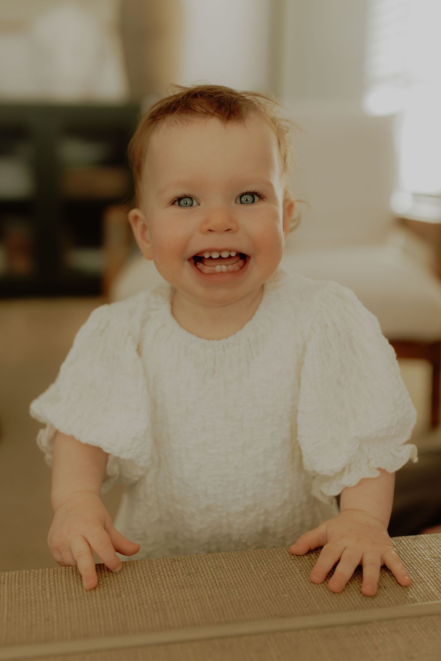 Smiling baby girl with blue eyes wearing a white textured dress, standing and leaning on a surface indoors.