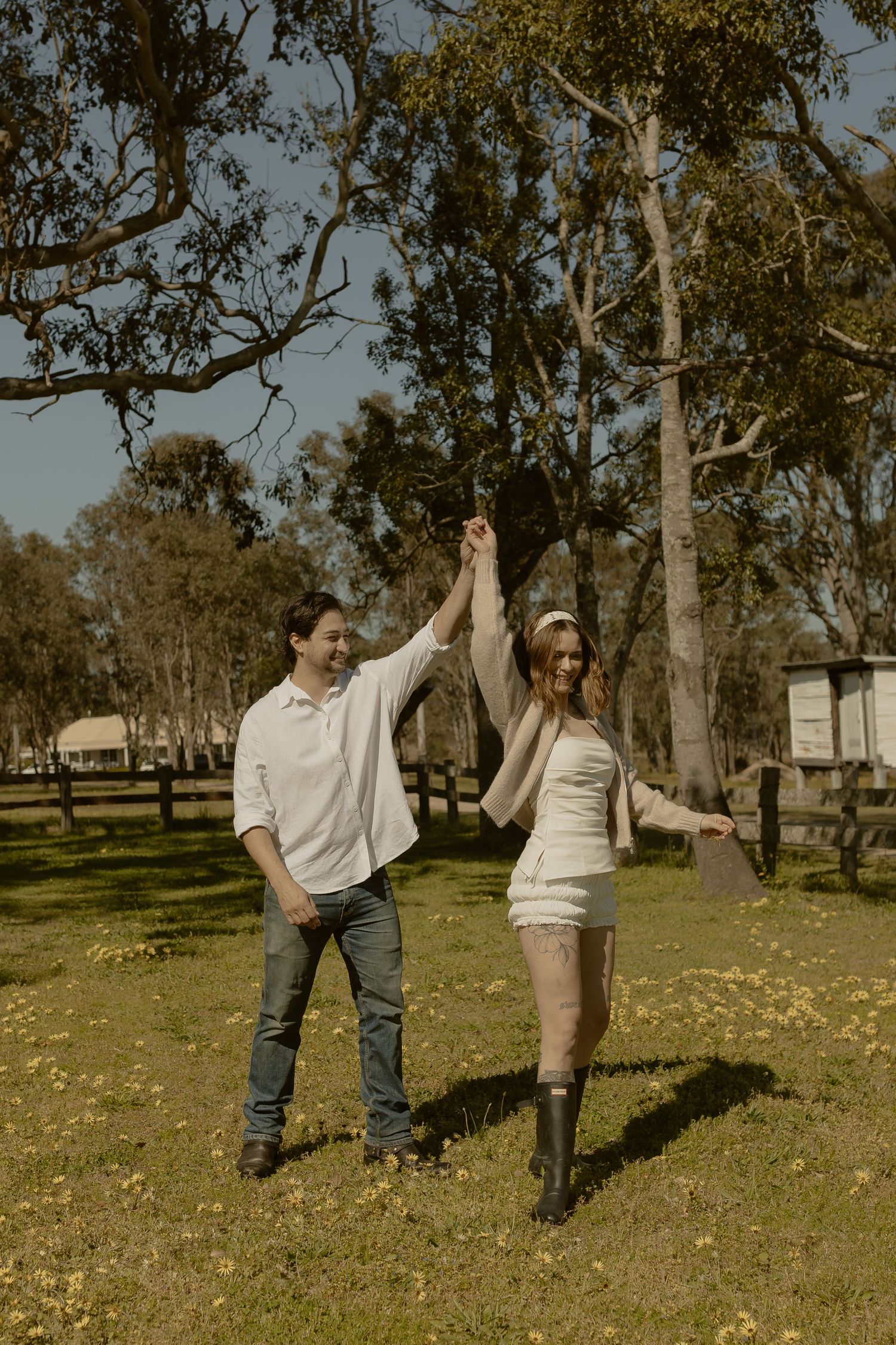 Young couple dancing and laughing together in a sunny park with scattered autumn leaves and tall trees.