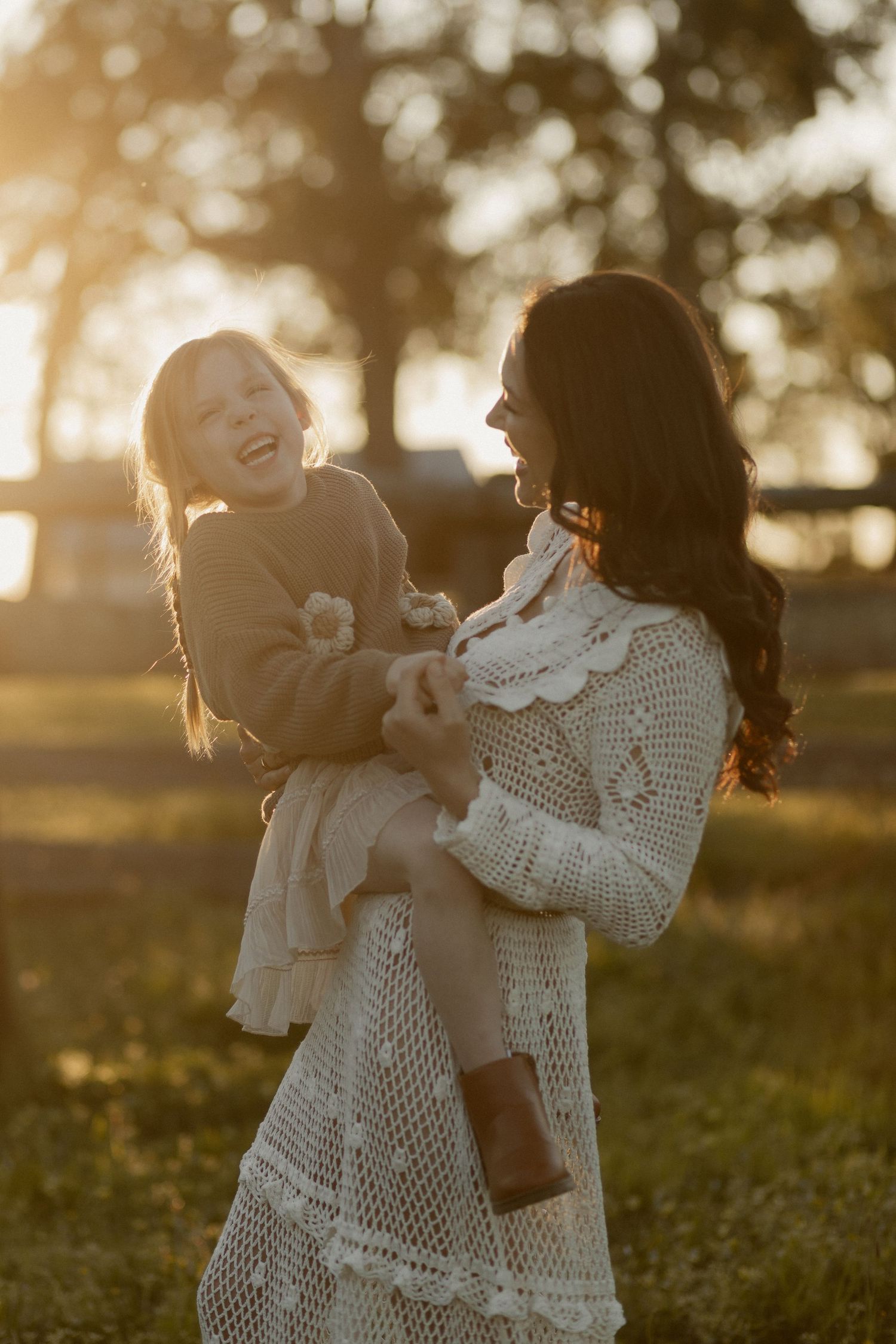 Two girls in white dresses laugh and embrace while playing in a sunlit field at golden hour.