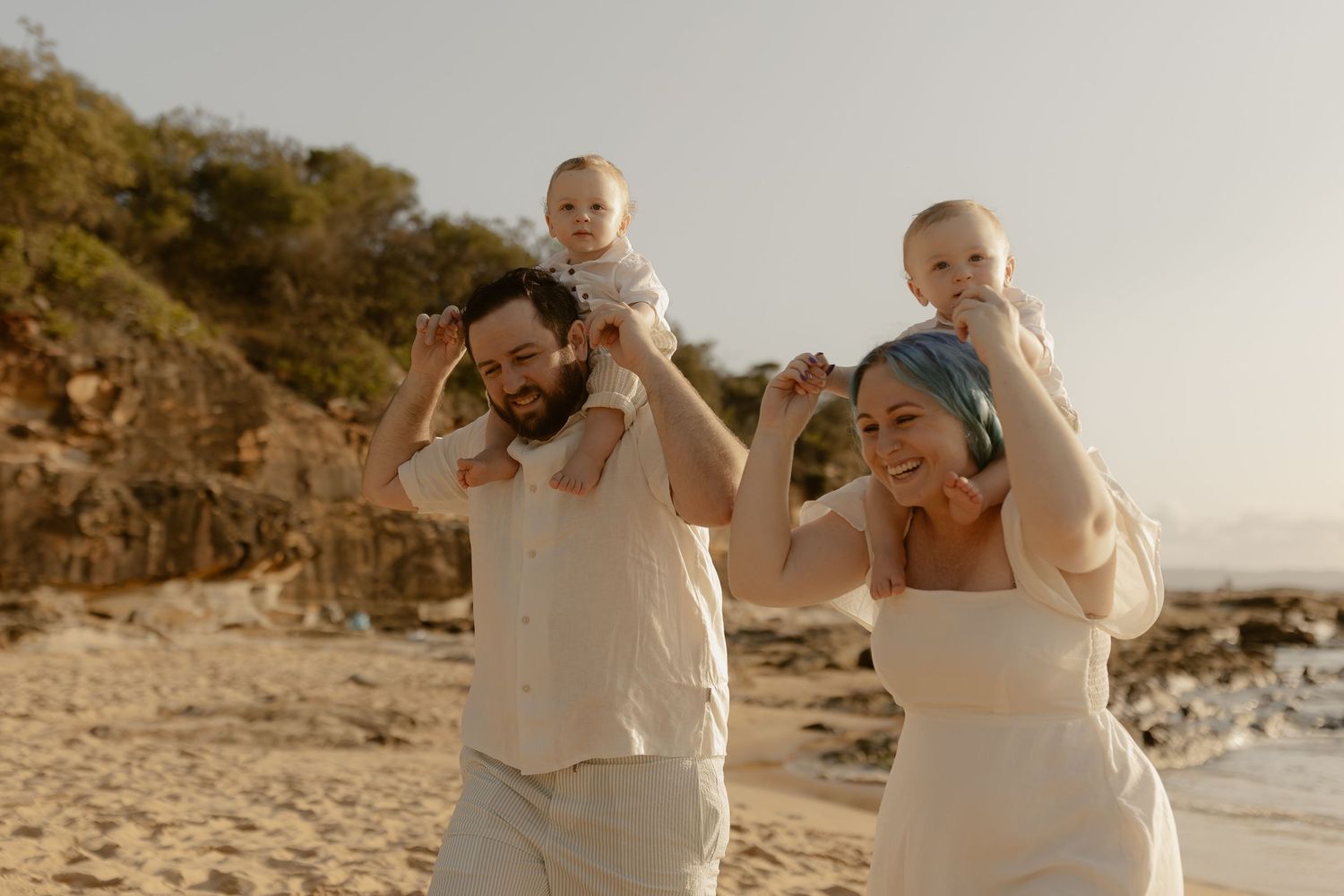 A joyful couple in white outfits celebrates their beach wedding with playful poses along a scenic coastal shoreline.