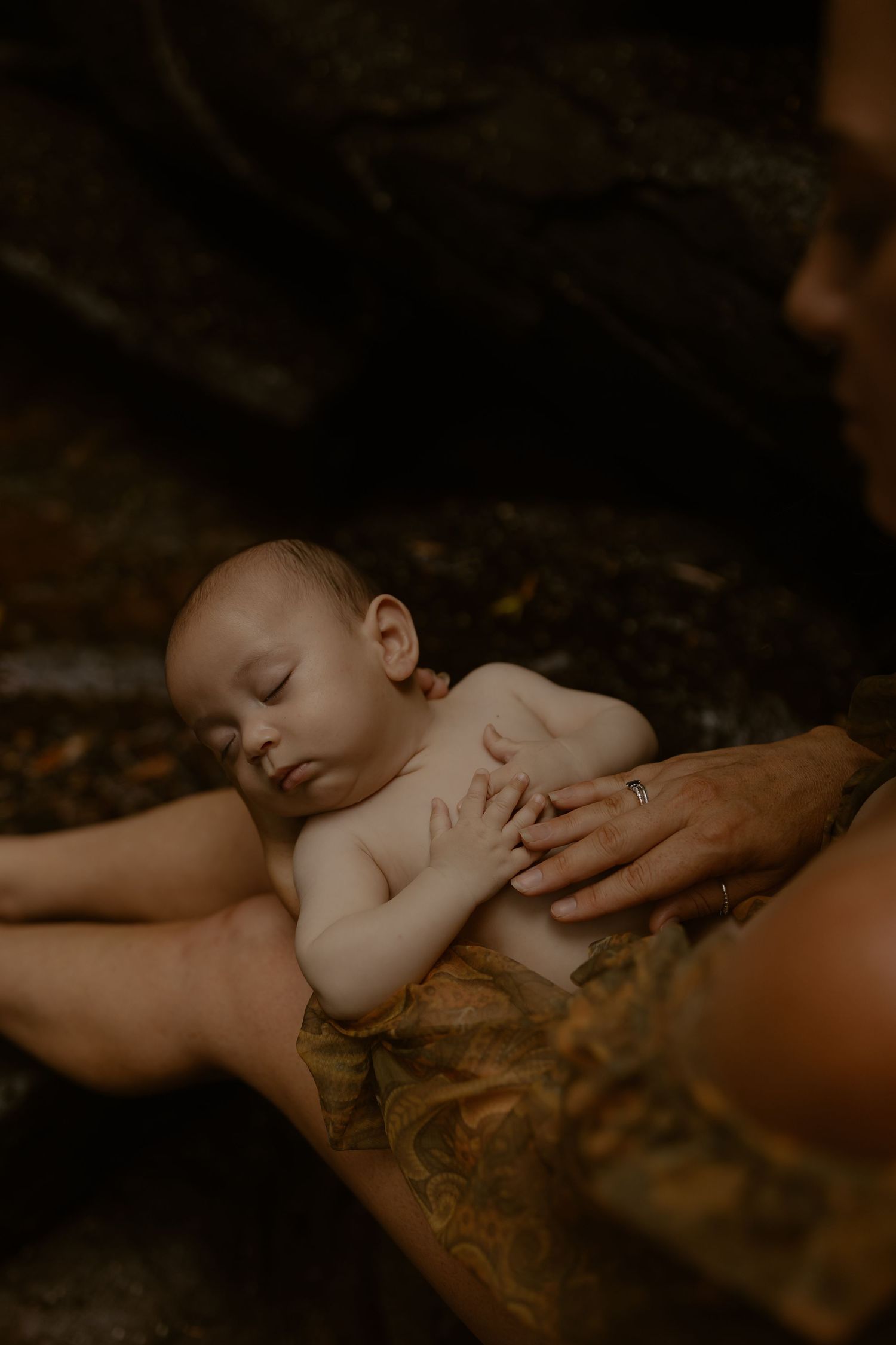A sleeping baby rests peacefully on a parent's lap while a gentle hand provides comfort in a moody, warm-toned setting.