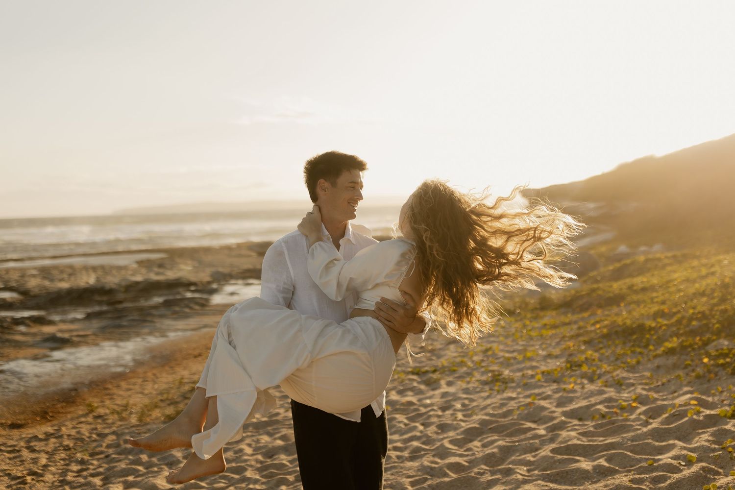 Couple in white clothing dancing on beach cliffs during sunset with ocean views.