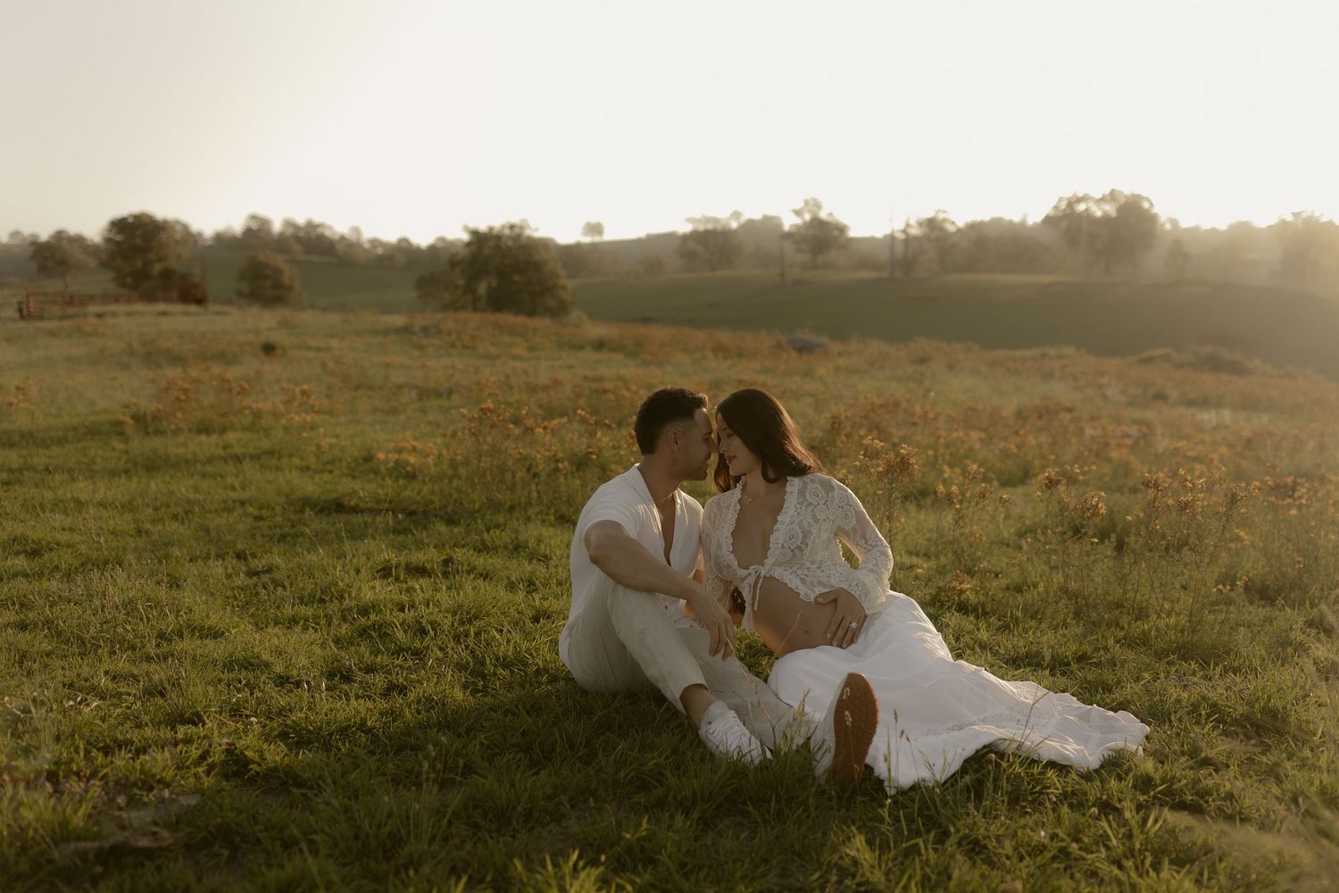 A romantic couple in white outfits sits intimately together on a grassy hilltop during golden hour sunset.