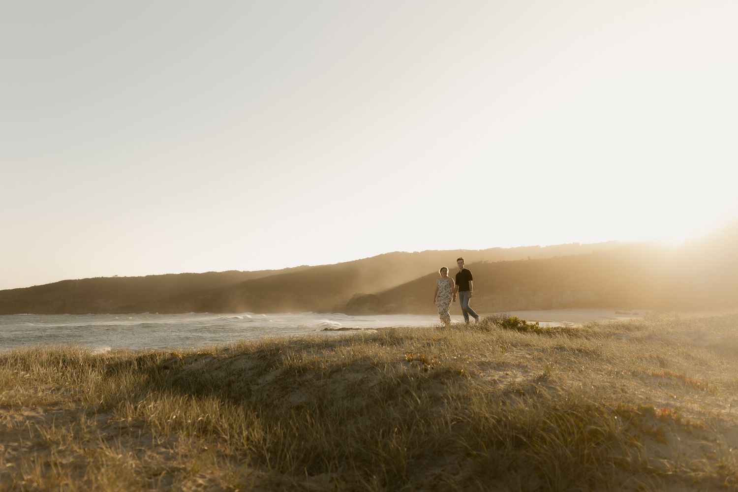 Couple walking along a misty coastal cliff at sunset with dramatic golden light.