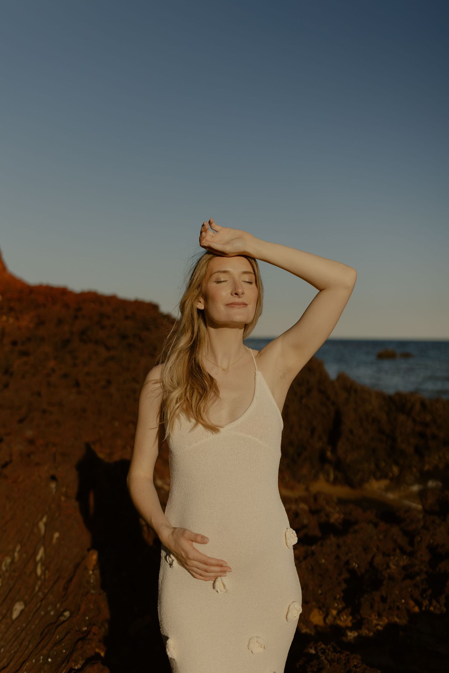 A serene maternity portrait at sunset shows a woman in a white dress posing against rocky coastal scenery.