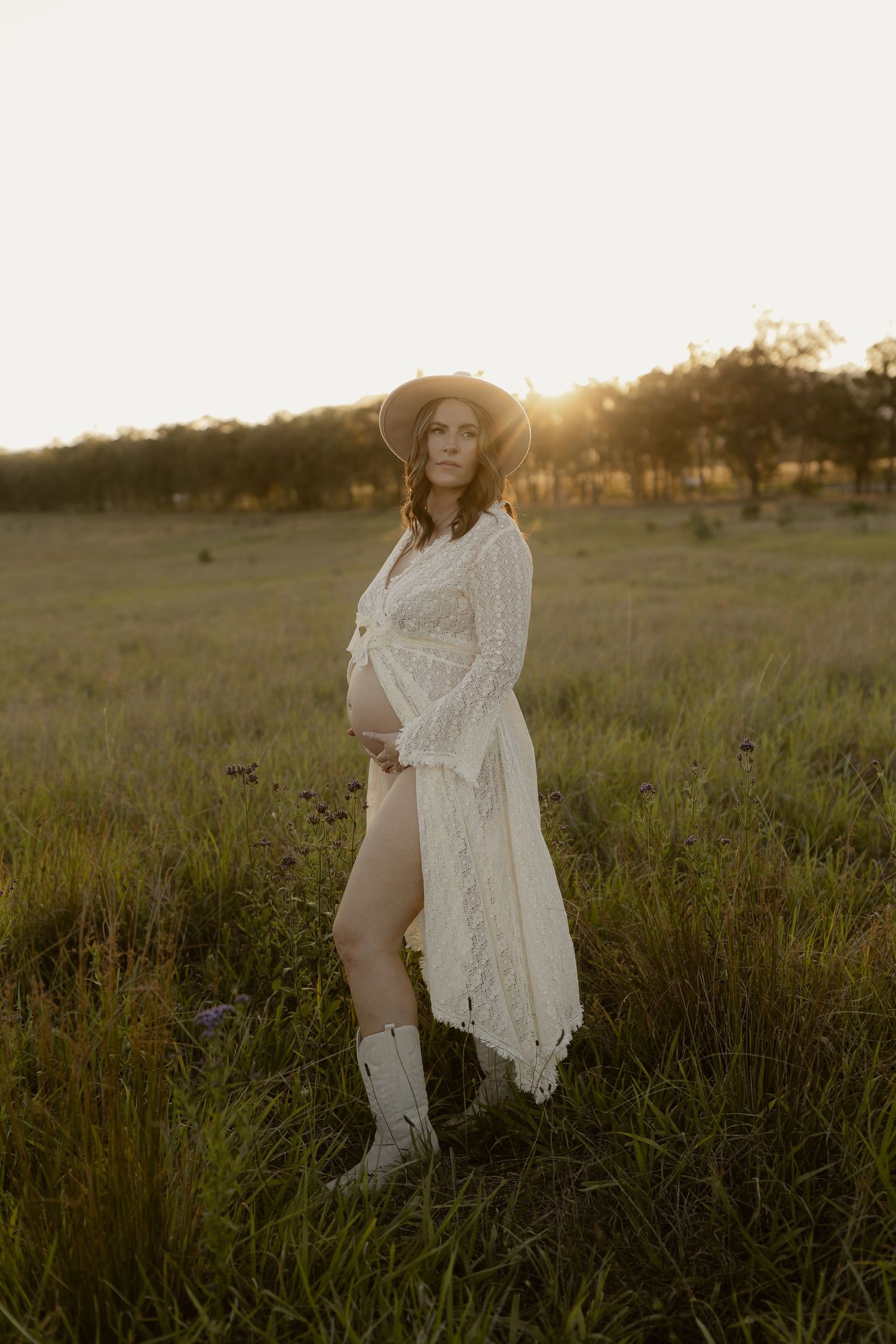 Figure in white lace dress and boots poses gracefully in tall grass during sunset.
