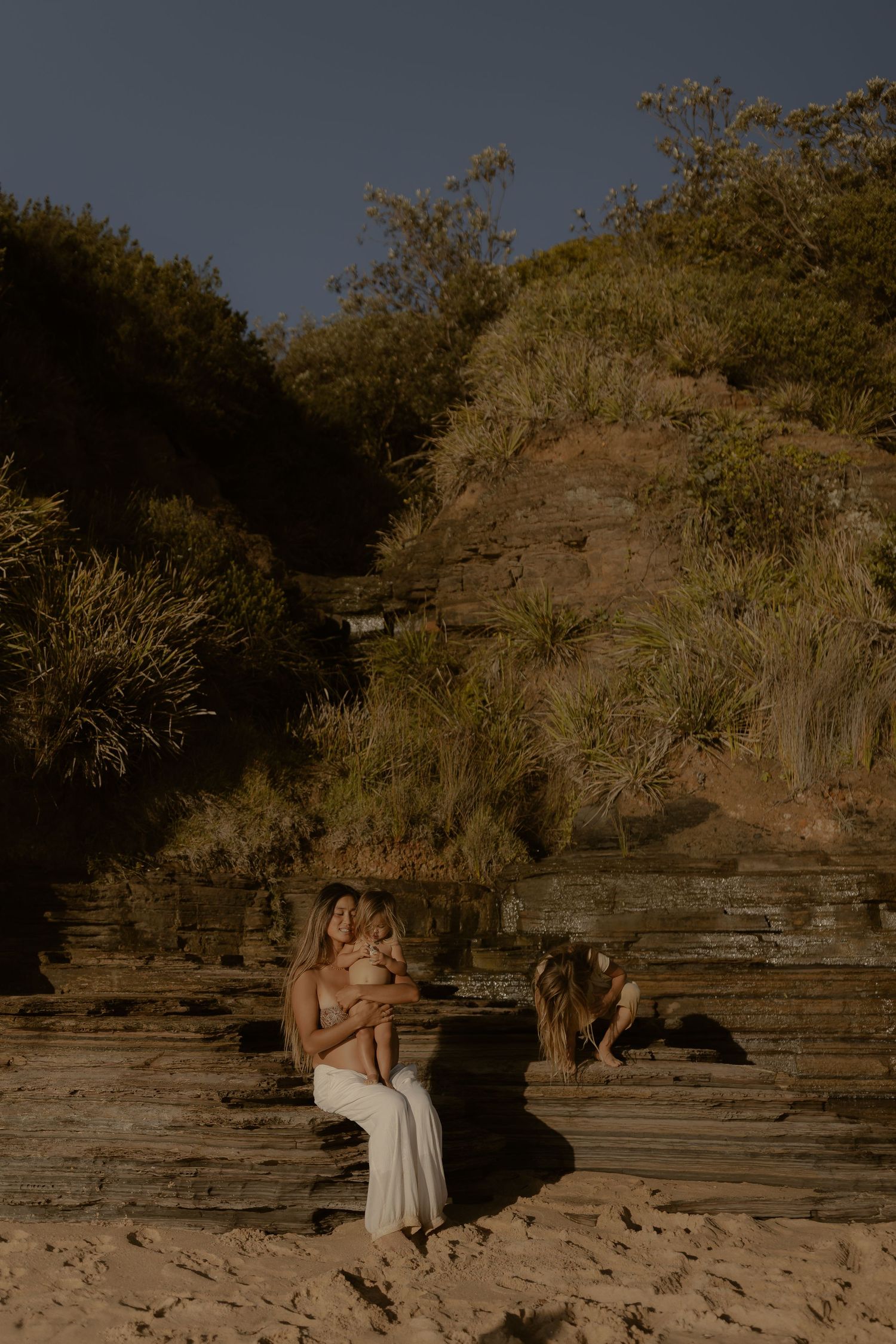 Two people in light dresses stand among rocky coastal terrain at sunset.