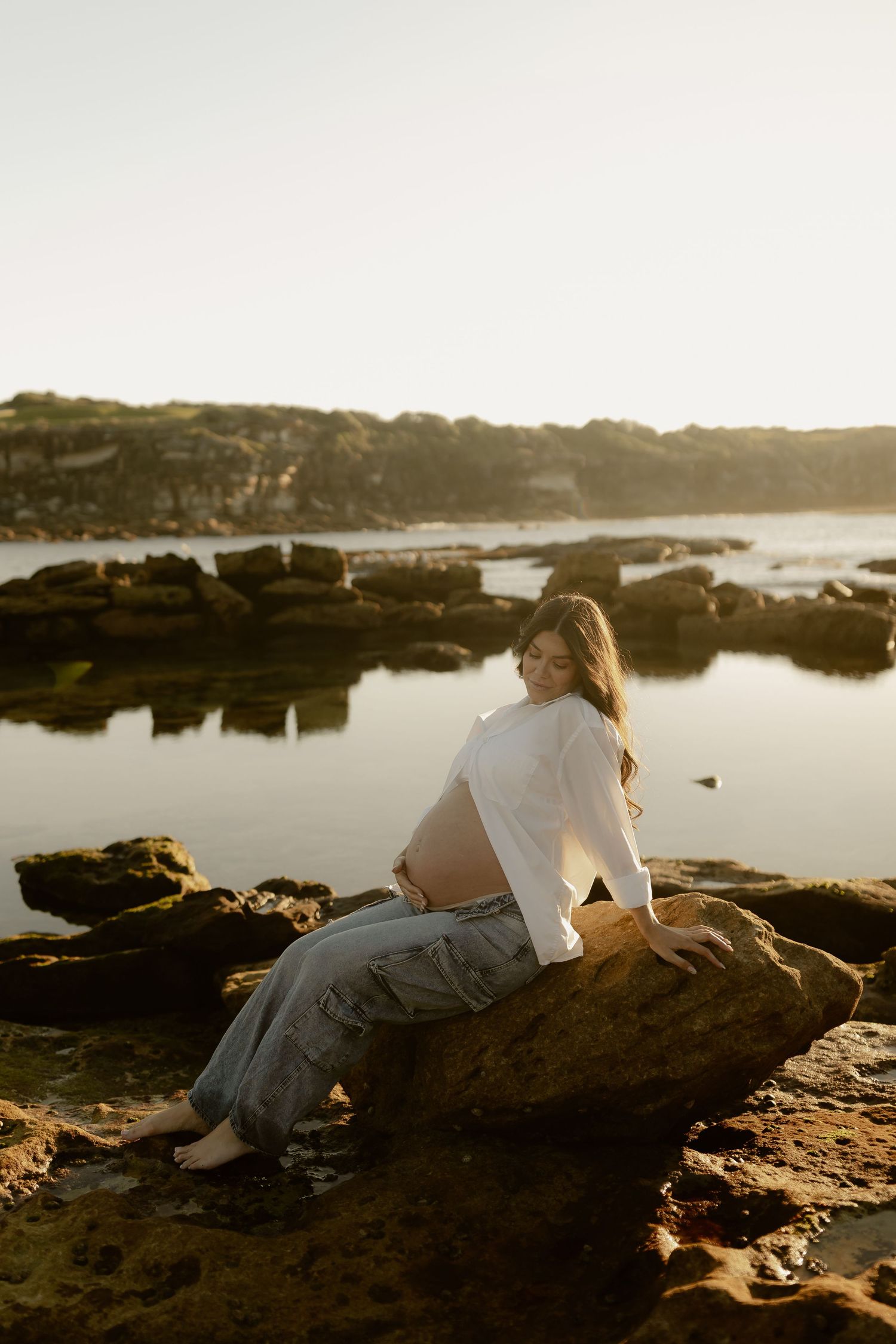 Smiling pregnant woman sitting on coastal rocks at sunset, cradling her baby bump in white shirt.