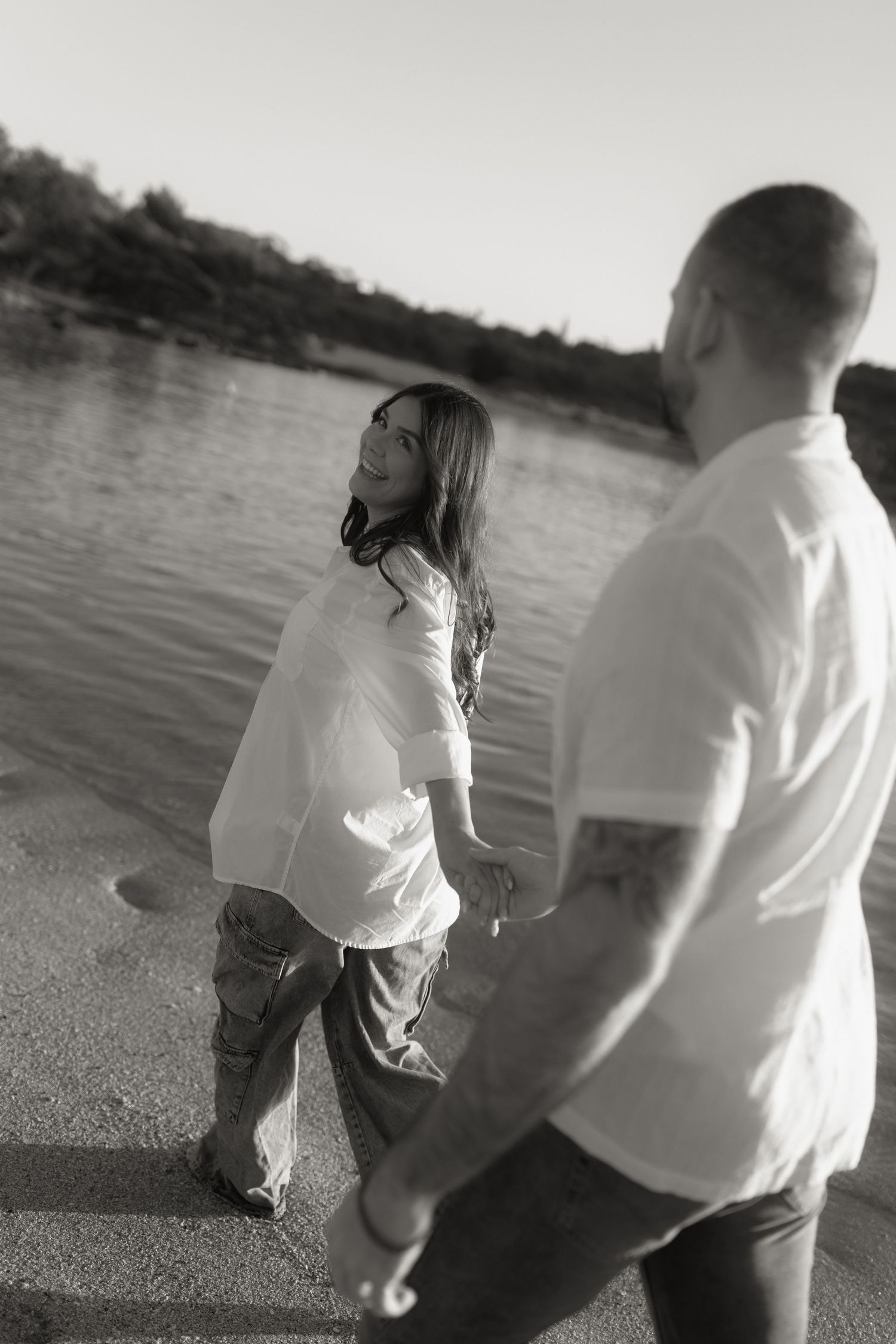 Black and white photo of pregnant couple holding hands and laughing near a sunlit river.