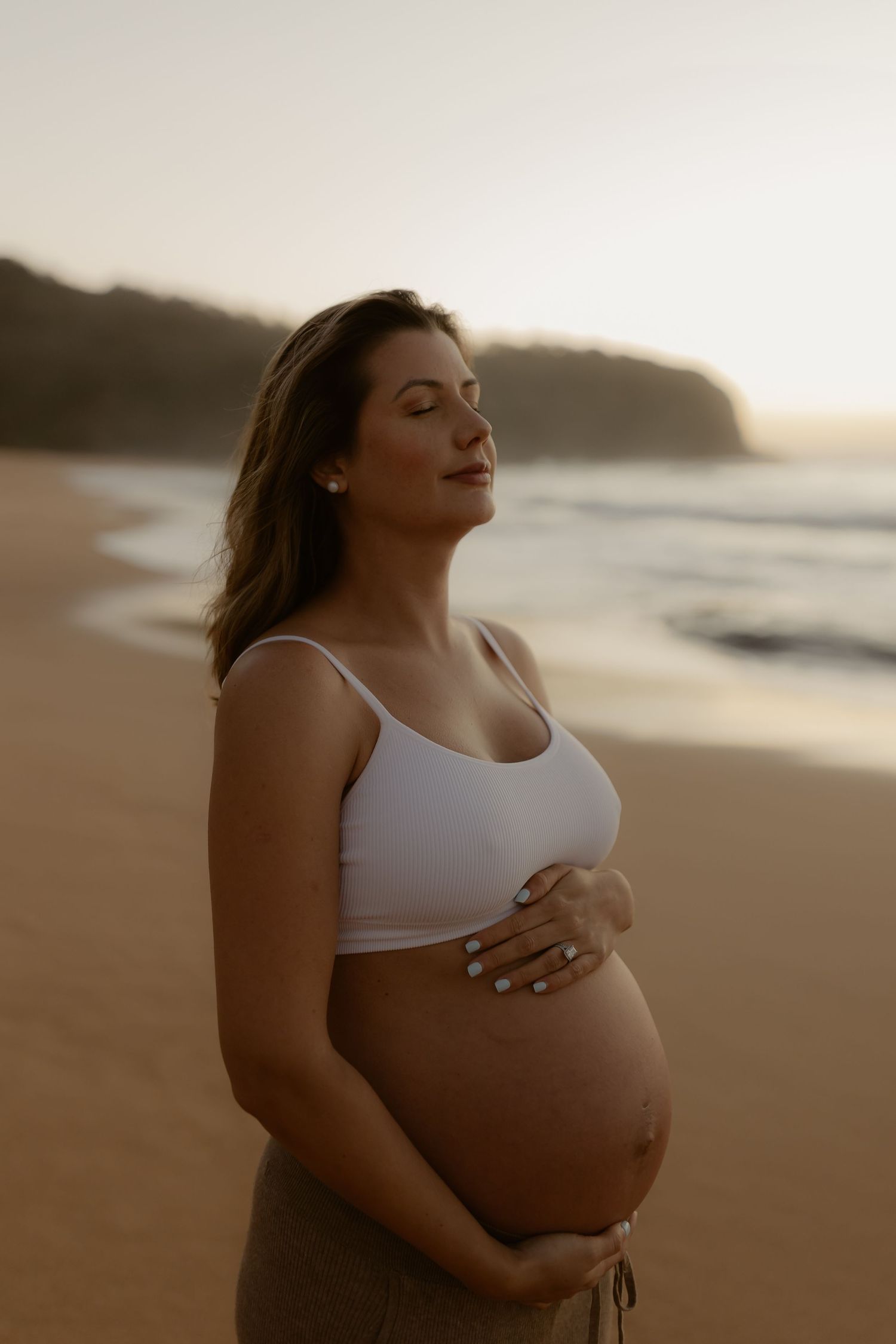 Pregnant woman in white crop top cradling her baby bump on a serene beach at golden hour sunset.