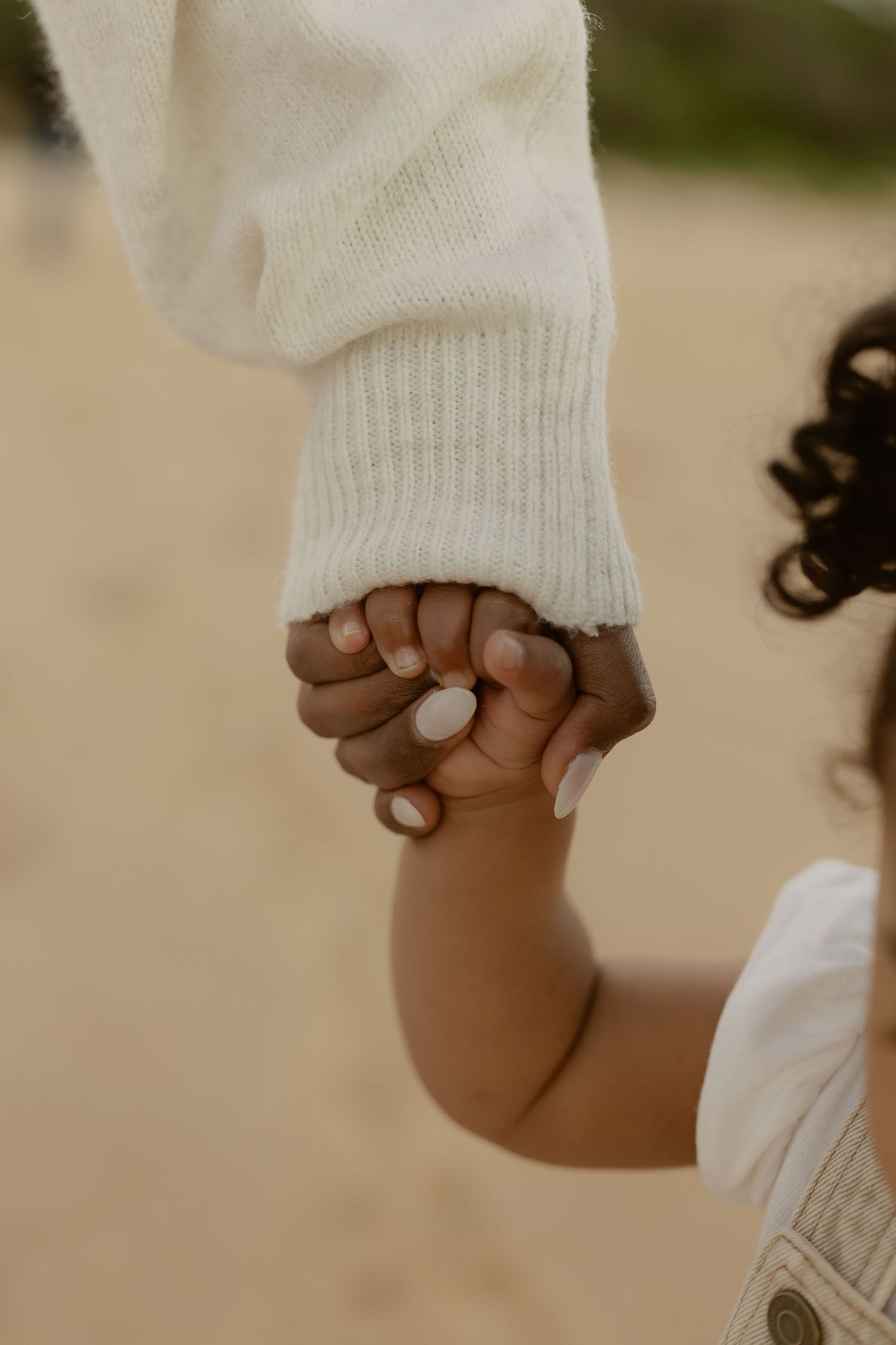 Adult and toddler holding hands outdoors, wearing white knit and denim overalls.