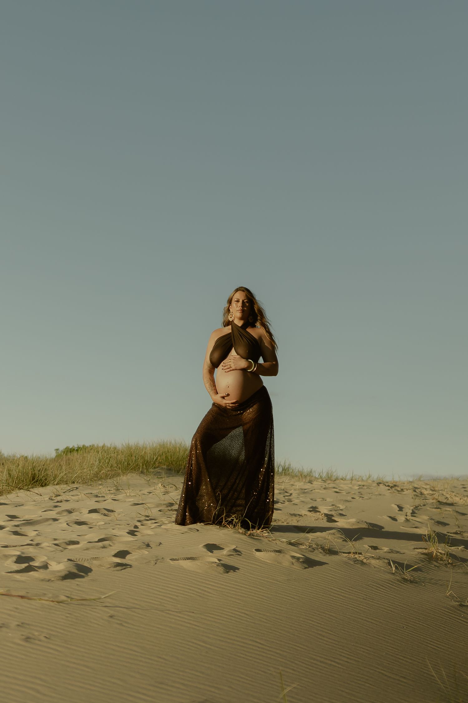Pregnant woman in sheer dark dress cradling baby bump on sandy dunes under clear blue sky.