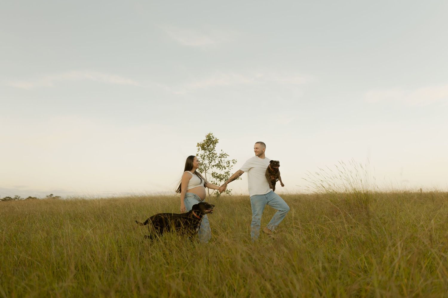 A couple and their black dog play in a lush green field under a soft sky at golden hour.