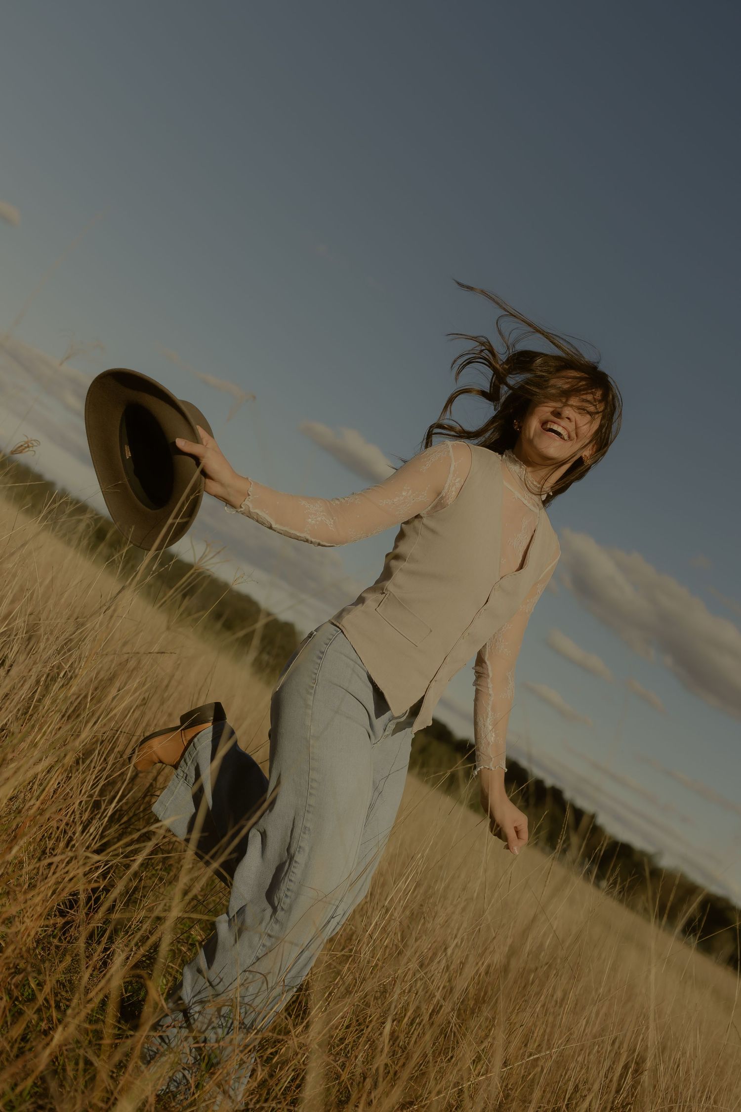 Joyful woman dancing in golden grass field, holding a hat with hair blowing in the wind under a blue sky.