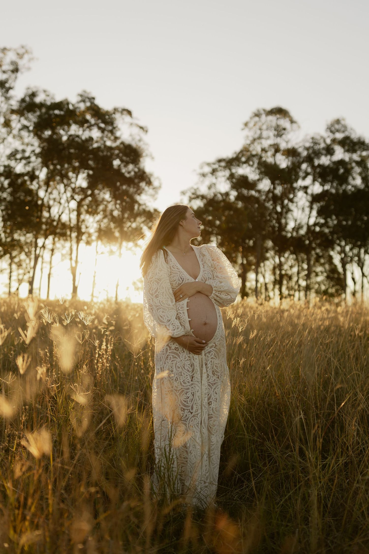 Pregnant woman in white lace dress holding baby bump in golden grass field at sunset.