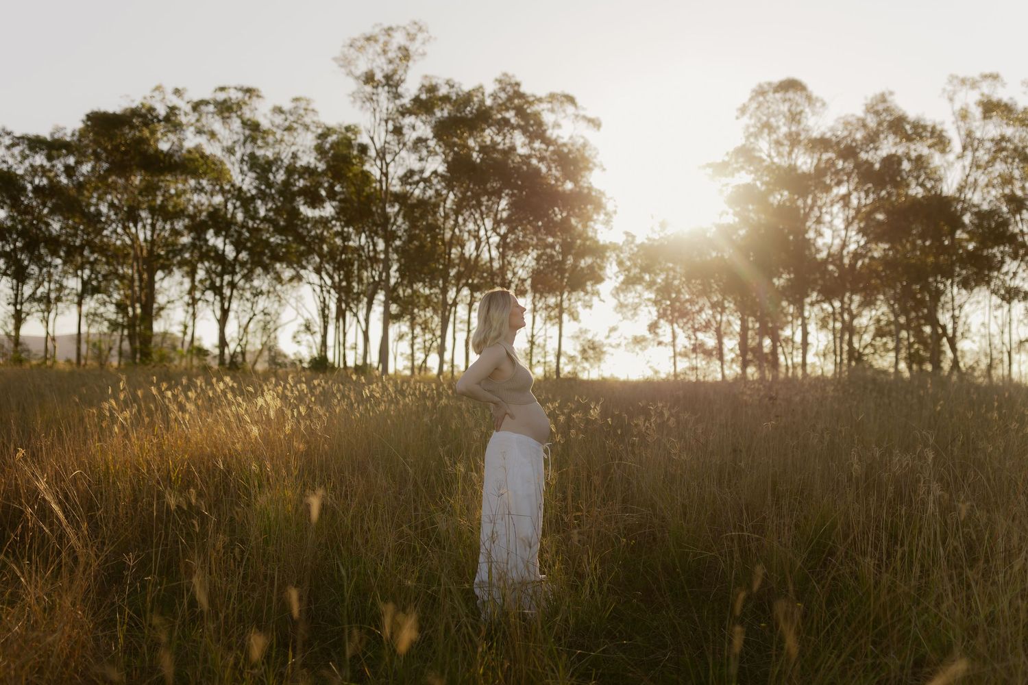 A woman in a white dress stands in golden tall grass at sunset with trees glowing in warm backlight.