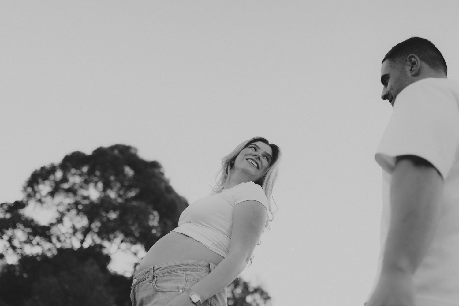 Black and white photo of a smiling pregnant woman with her partner outdoors near trees.