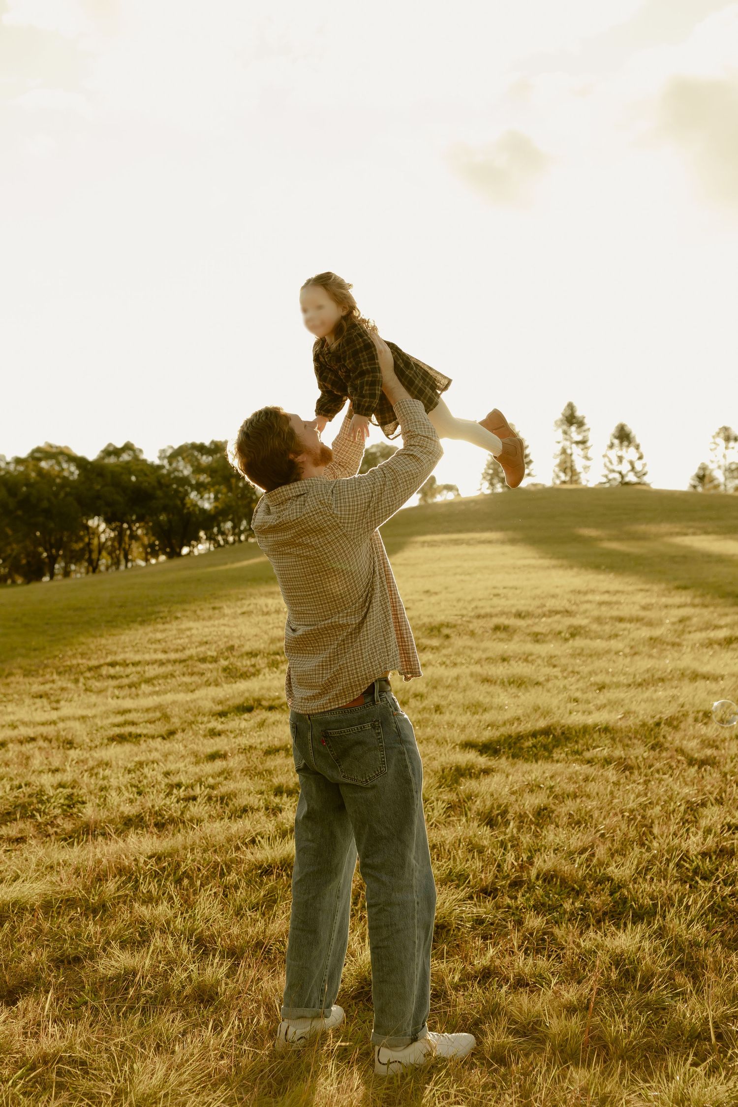 A father lifts his young daughter high in the air on a sunny grassy hillside at golden hour.