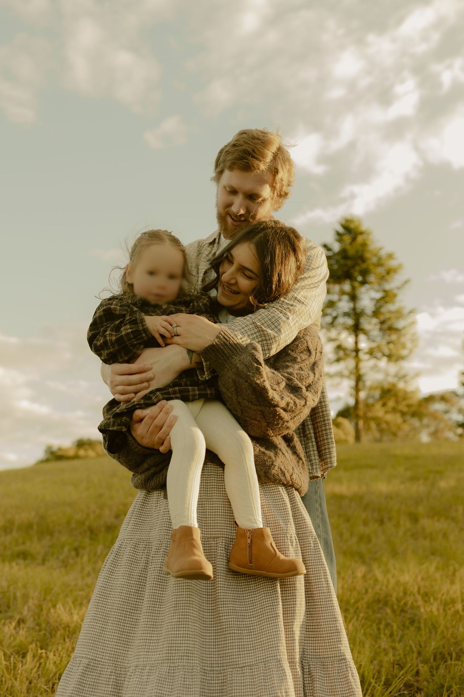 A happy family of three sharing a warm embrace in a green meadow on a cloudy day.