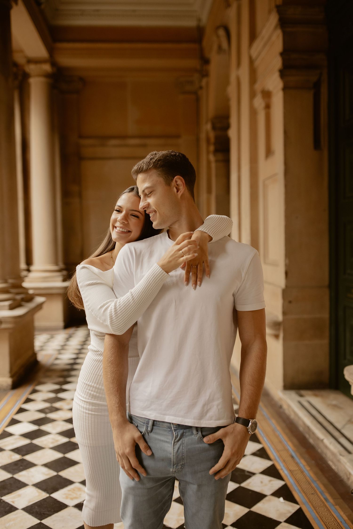 Romantic couple in white outfits embracing in an elegant hallway with checkered floor and classical columns.