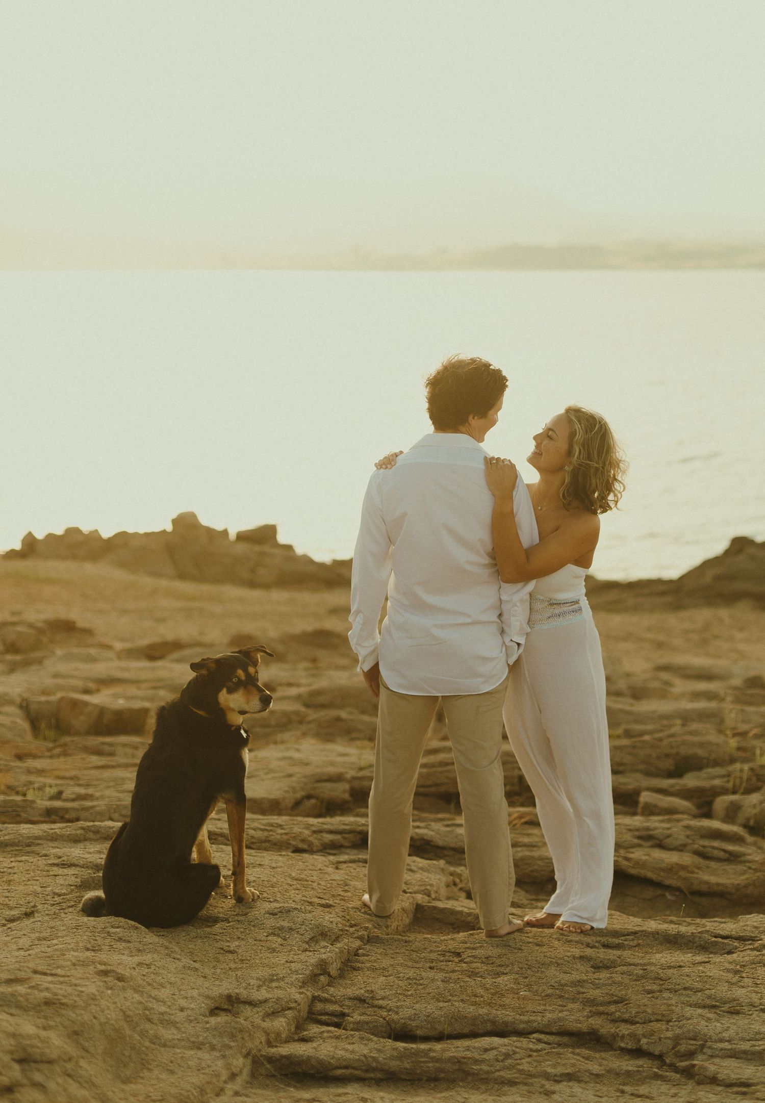 Couple shares romantic moment on rocky beach at sunset with their black dog sitting beside them.