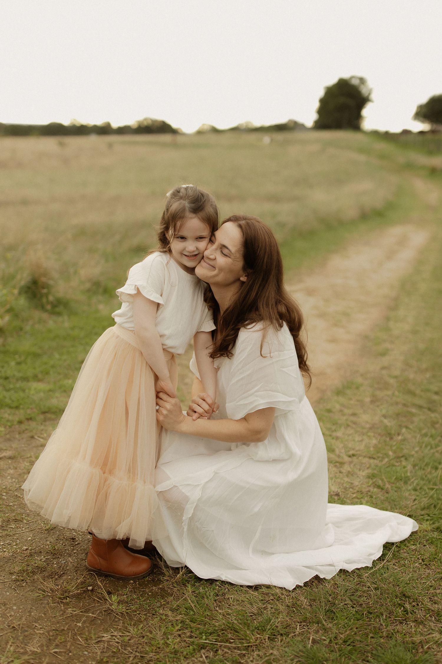 Two figures in white dresses share tender moments on a dirt path through a grassy hillside.