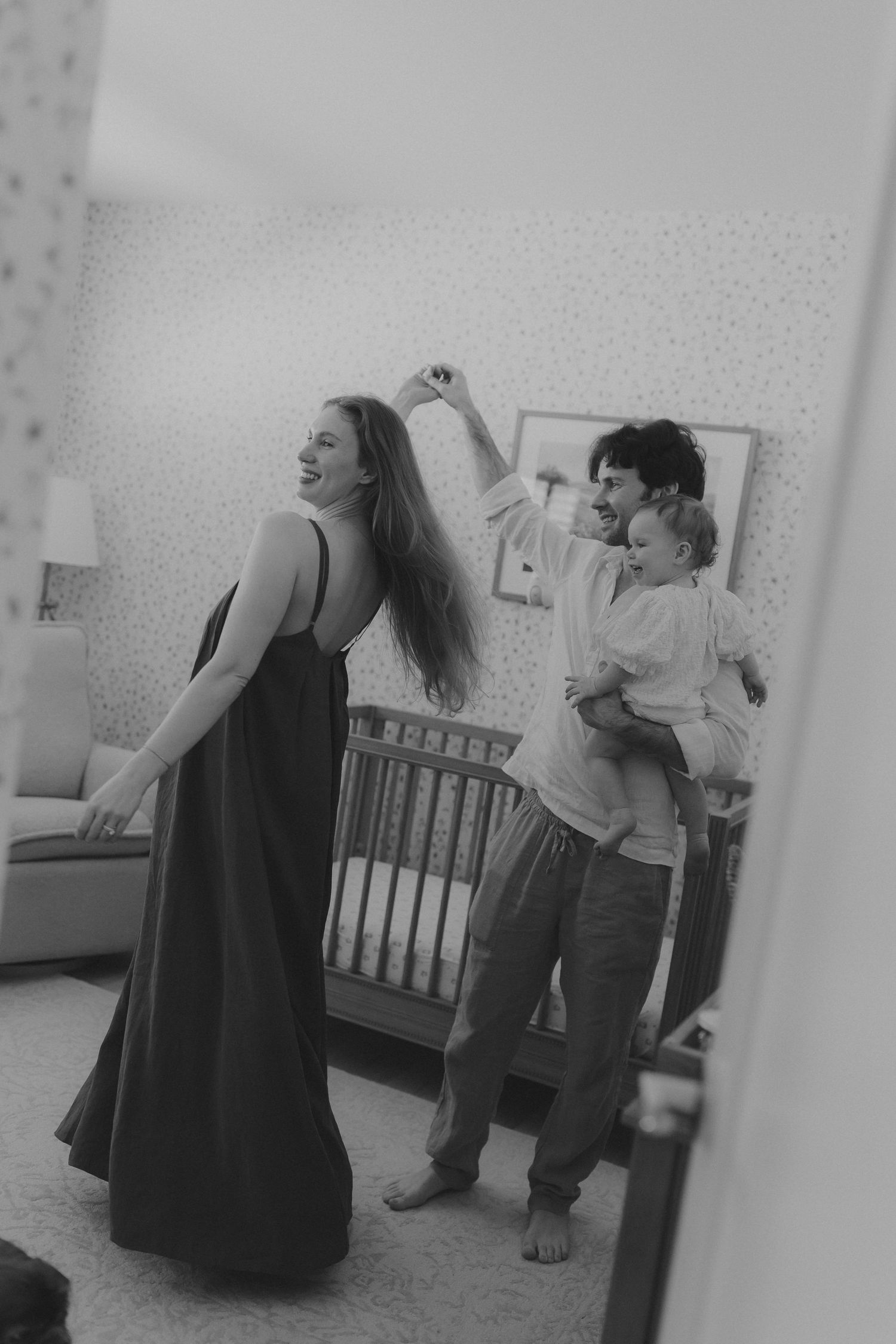 Black and white photo of a joyful family dancing in a nursery, mother in maxi dress, father holding baby.