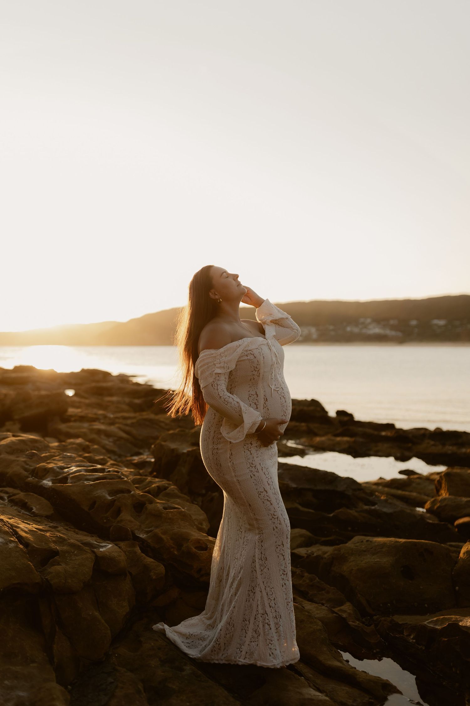 Bride in flowing white dress stands on rocky coastline during golden hour sunset.