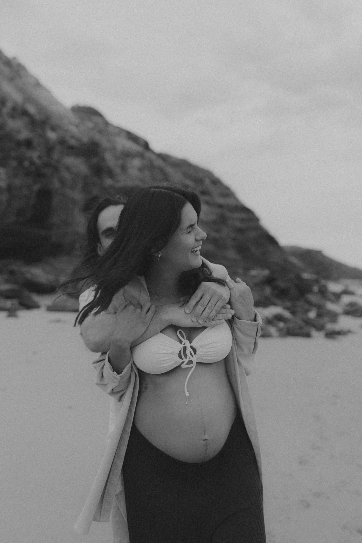 An expectant mother in a flowing coat embraces her belly while standing on a rocky beach.