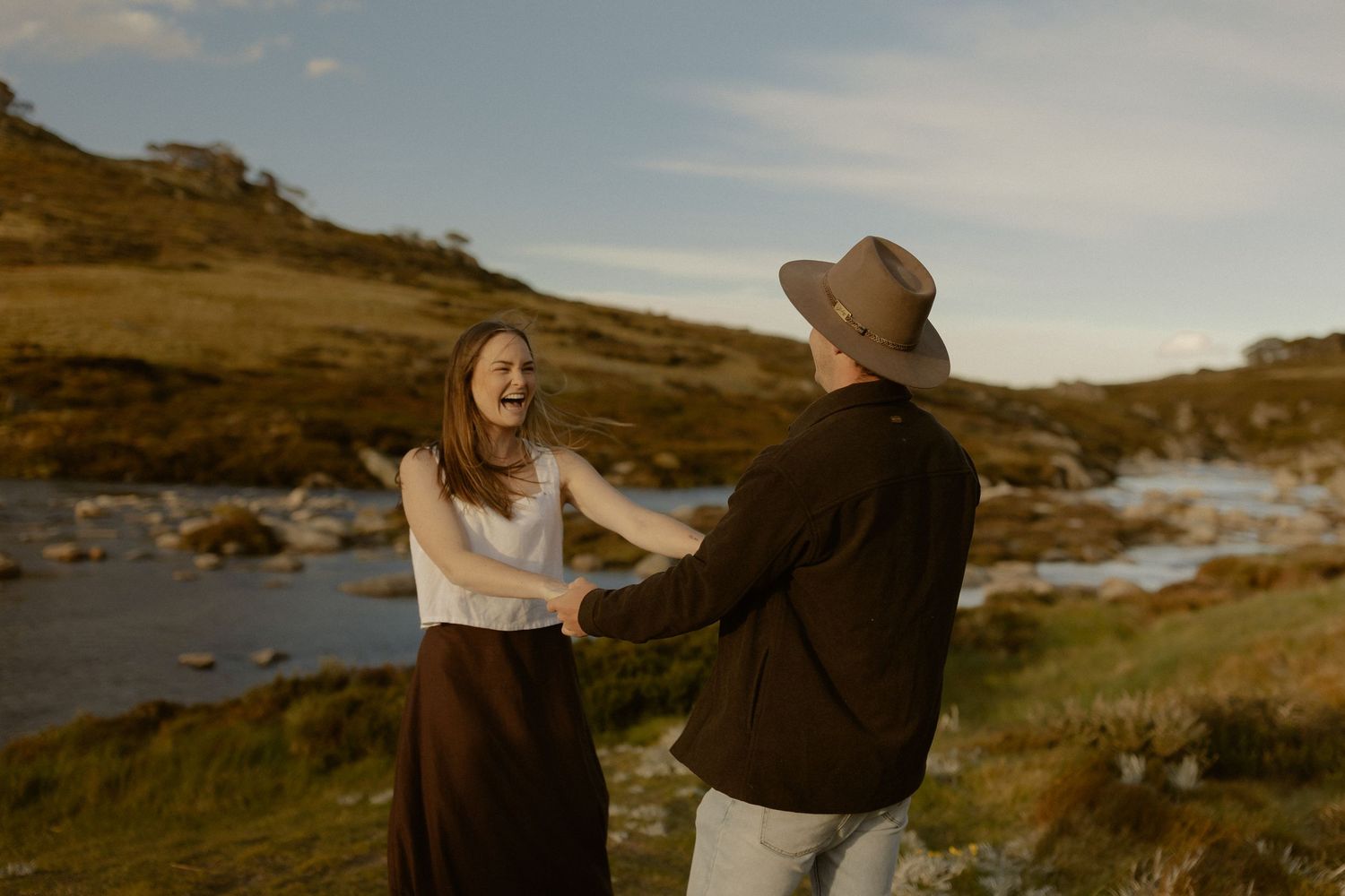 Two people in warm clothing walk and embrace along a scenic coastal path with rolling hills and ocean views at sunset.