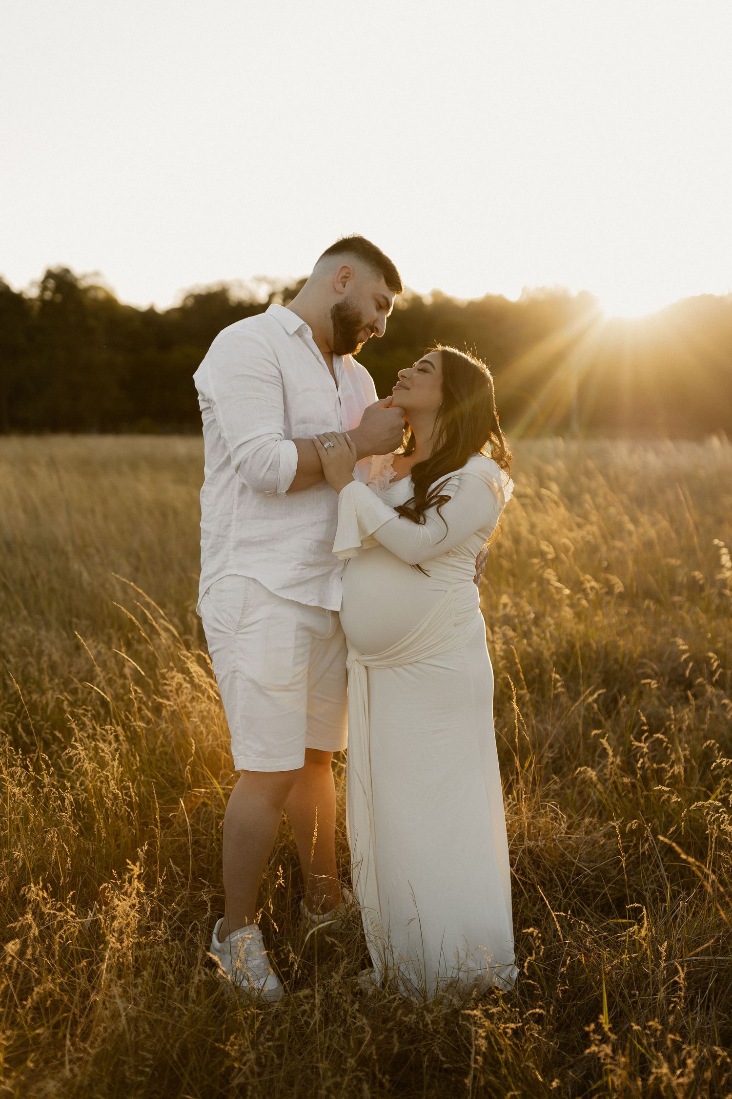 A romantic maternity photoshoot in a golden field at sunset with couple in white outfits embracing.