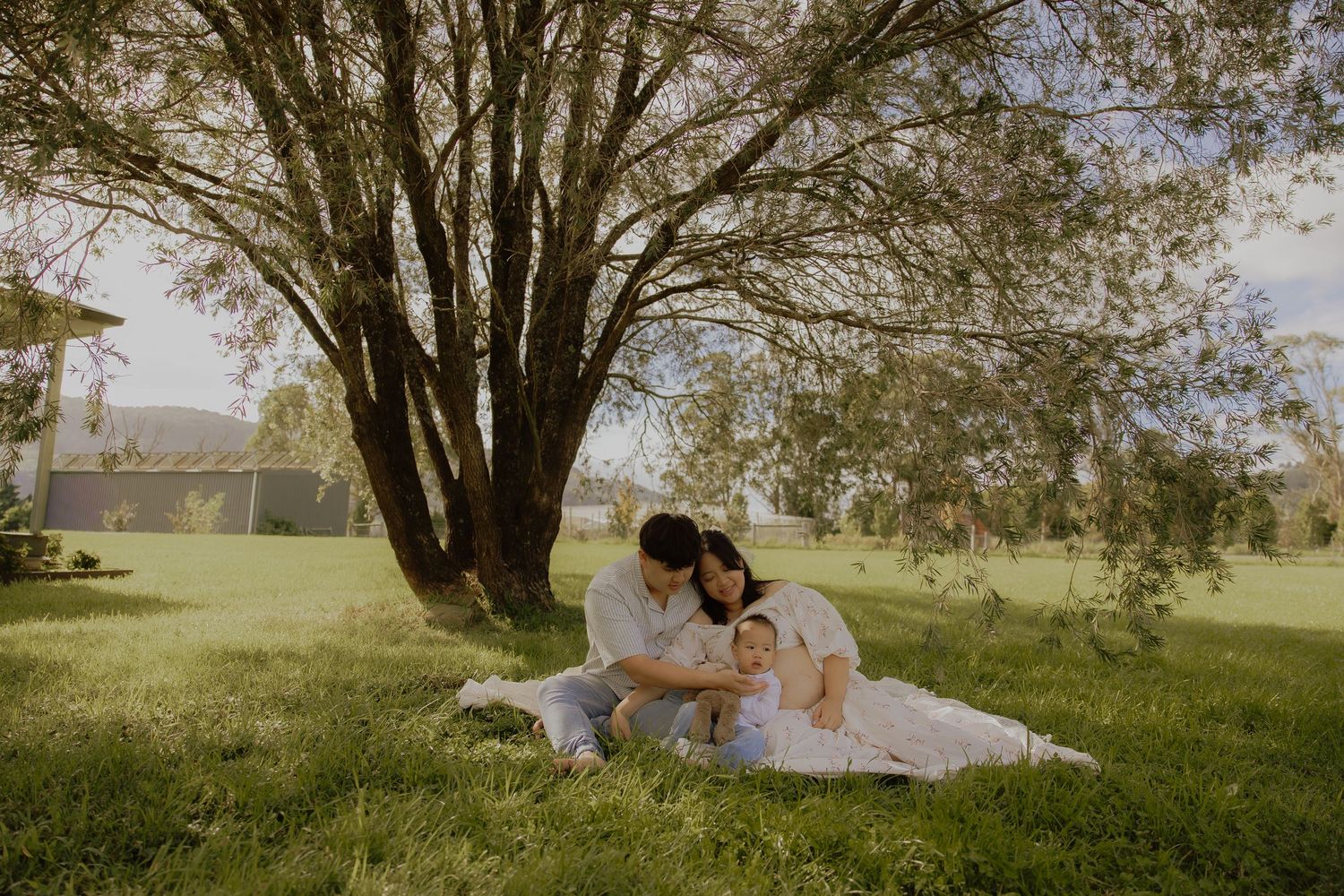 Family sitting together on a blanket under a large tree in a lush green park on a sunny day.