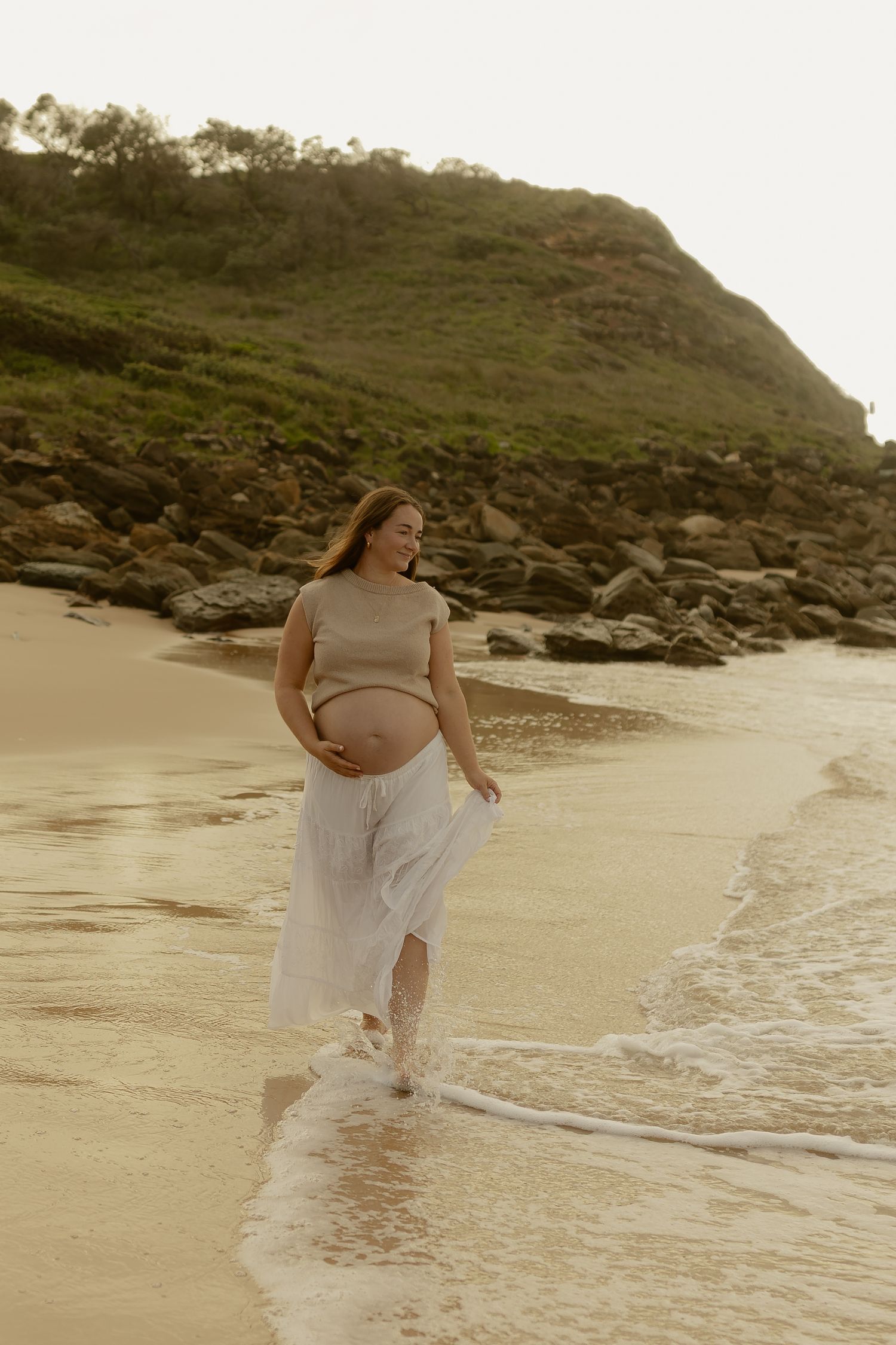A woman in flowing white dress dances joyfully at the water's edge on a secluded beach.